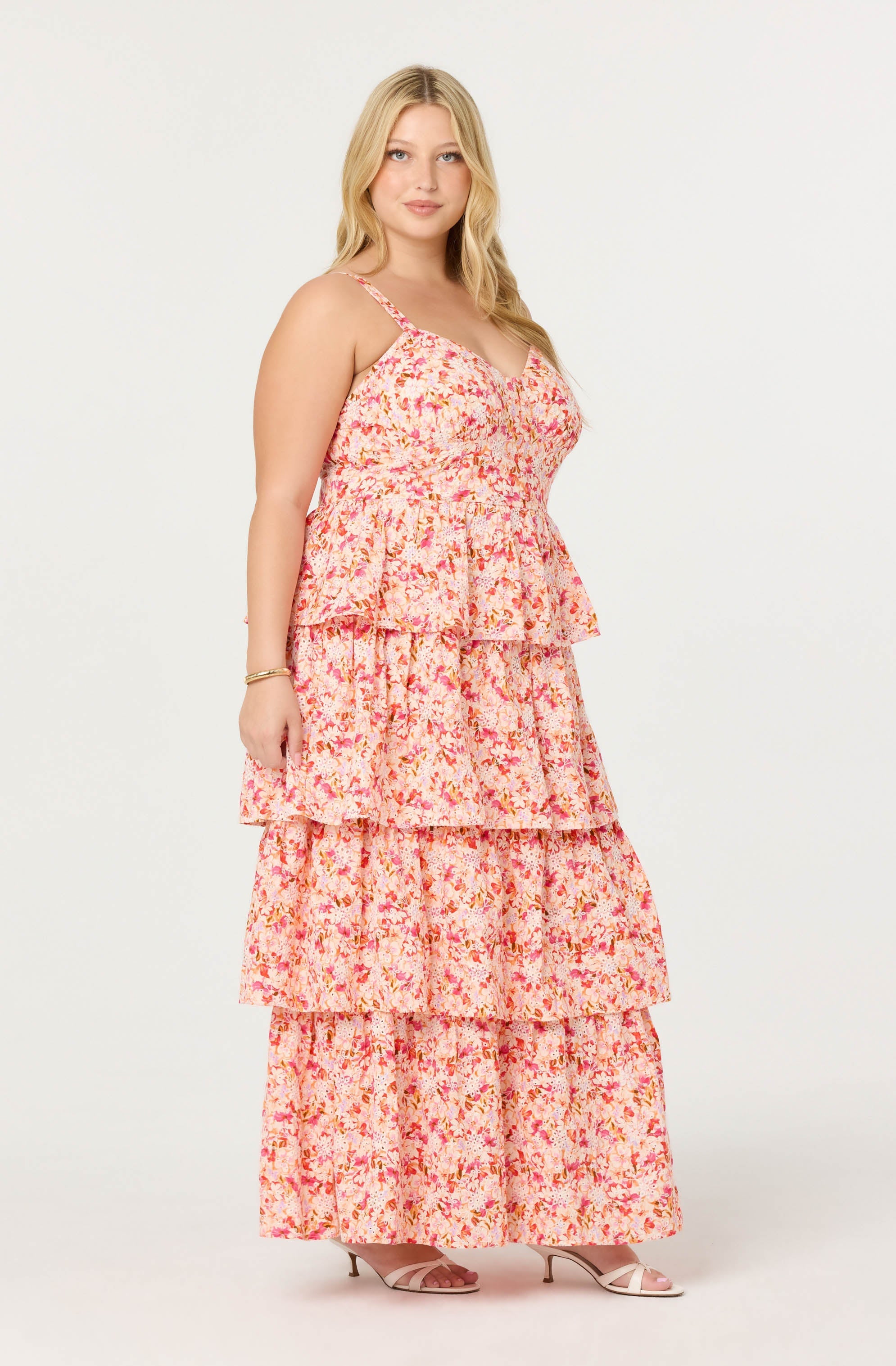 A woman wearing a pink and orange floral patterned maxi dress with tiered ruffle layers, spaghetti straps, and a fitted bodice. She is also wearing nude high-heeled sandals.