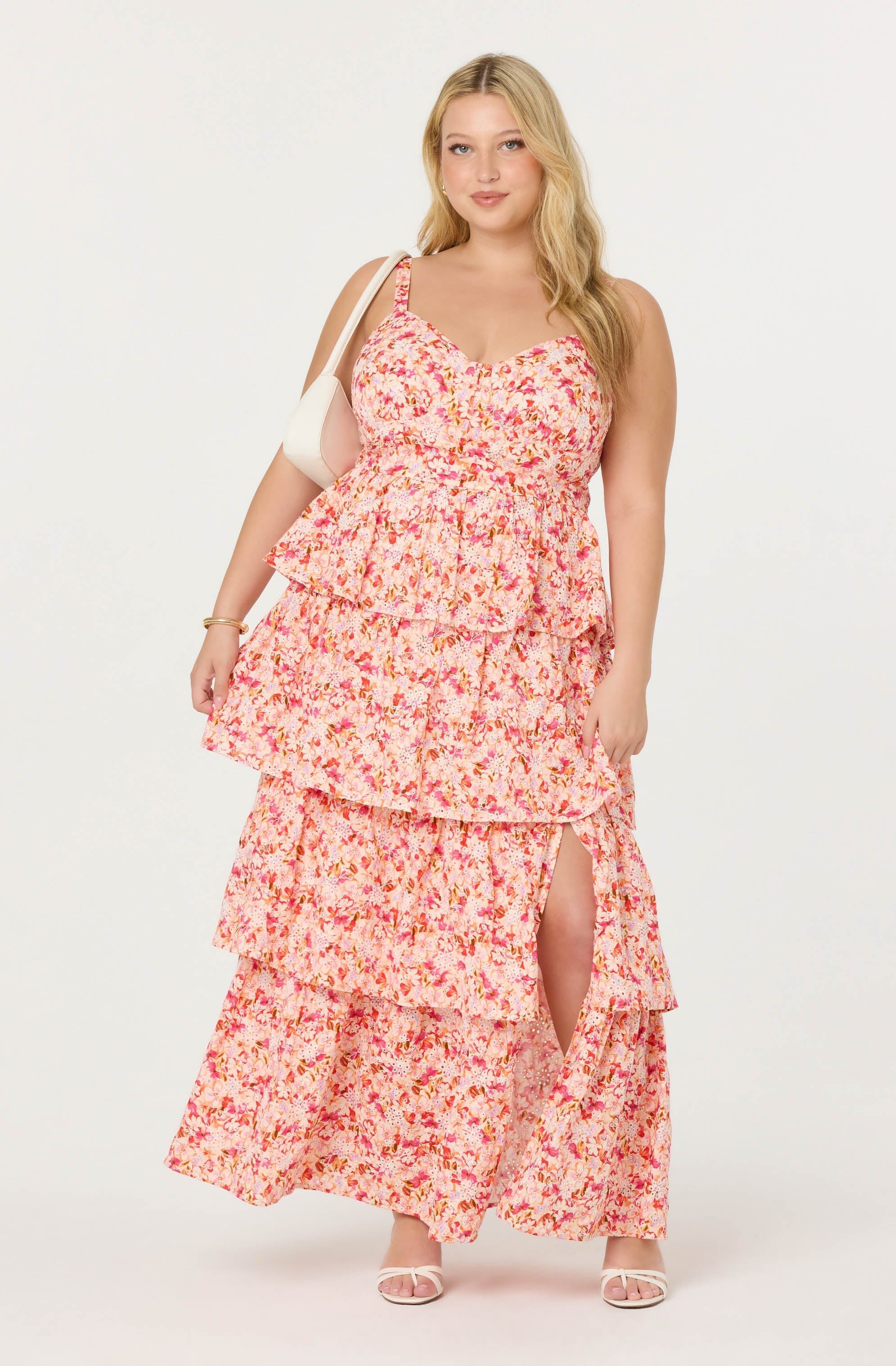 A woman is wearing a sleeveless, floral maxi dress with multiple ruffled tiers. The dress features a pink, orange, and cream-colored floral pattern, with a slit on one side revealing her leg. She is accessorized with a white handbag over her shoulder, a bracelet, and white open-toe heels.
