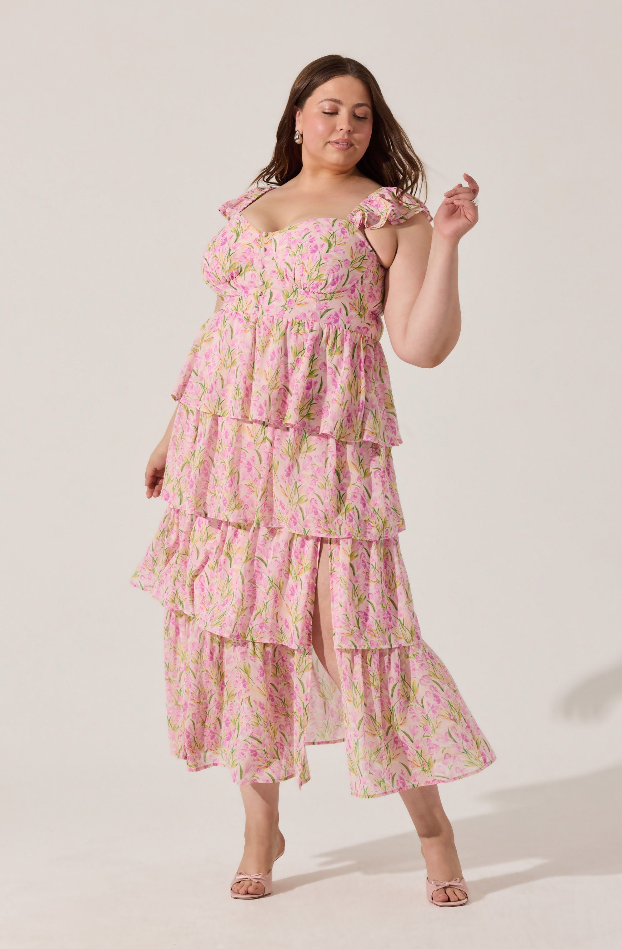 A woman is wearing a pink, floral-patterned dress with green and yellow accents. The dress features a fitted bodice with ruffled shoulder straps and an A-line skirt with multiple tiers of ruffles, creating a layered look. She is also wearing nude-colored open-toe heels.