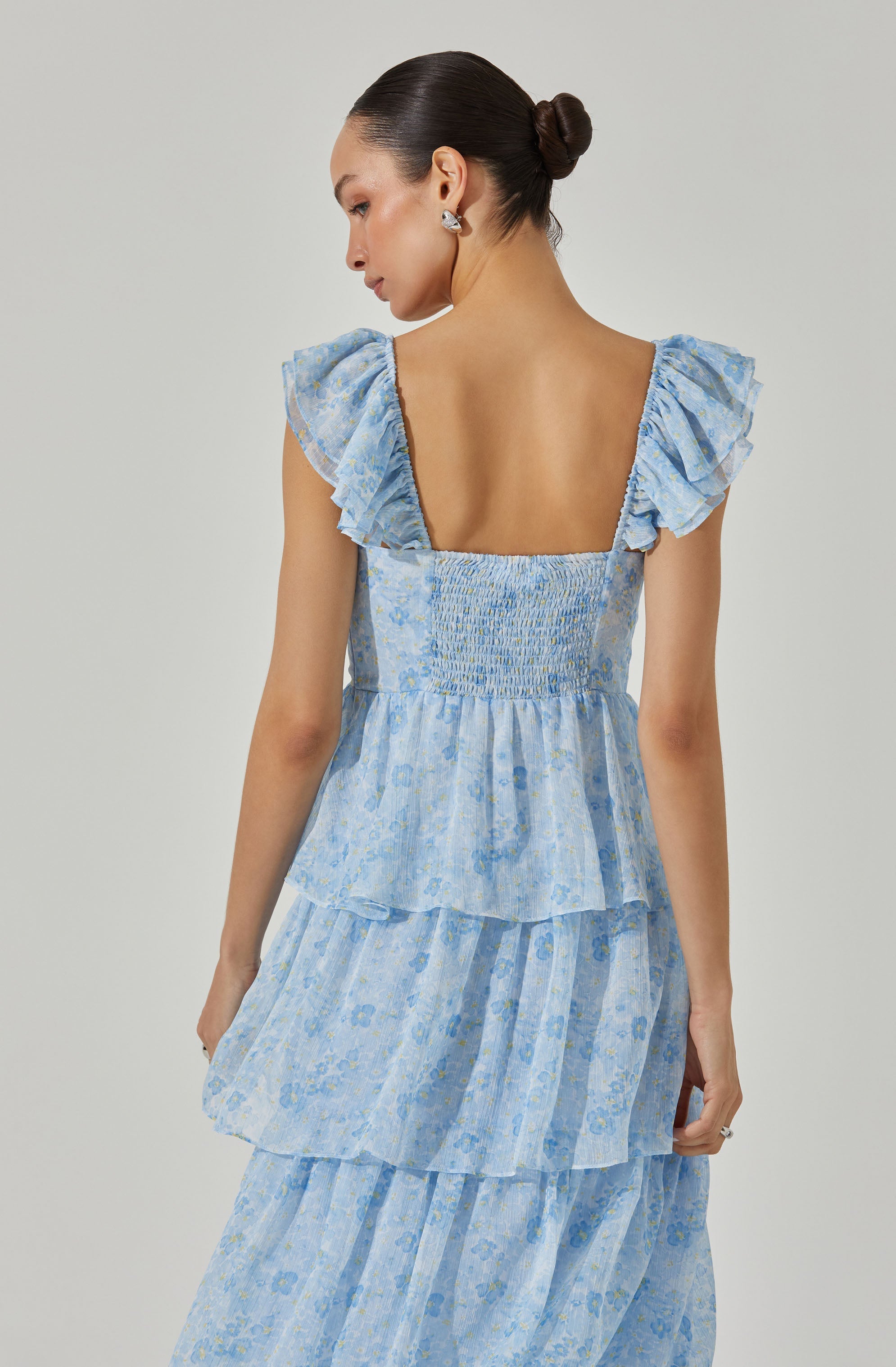 A woman is wearing a light blue, floral-printed dress with ruffled cap sleeves. The dress features smocked detailing at the bodice and multiple tiered layers in a flowing style. The fabric has a subtle floral pattern in shades of blue and yellow.