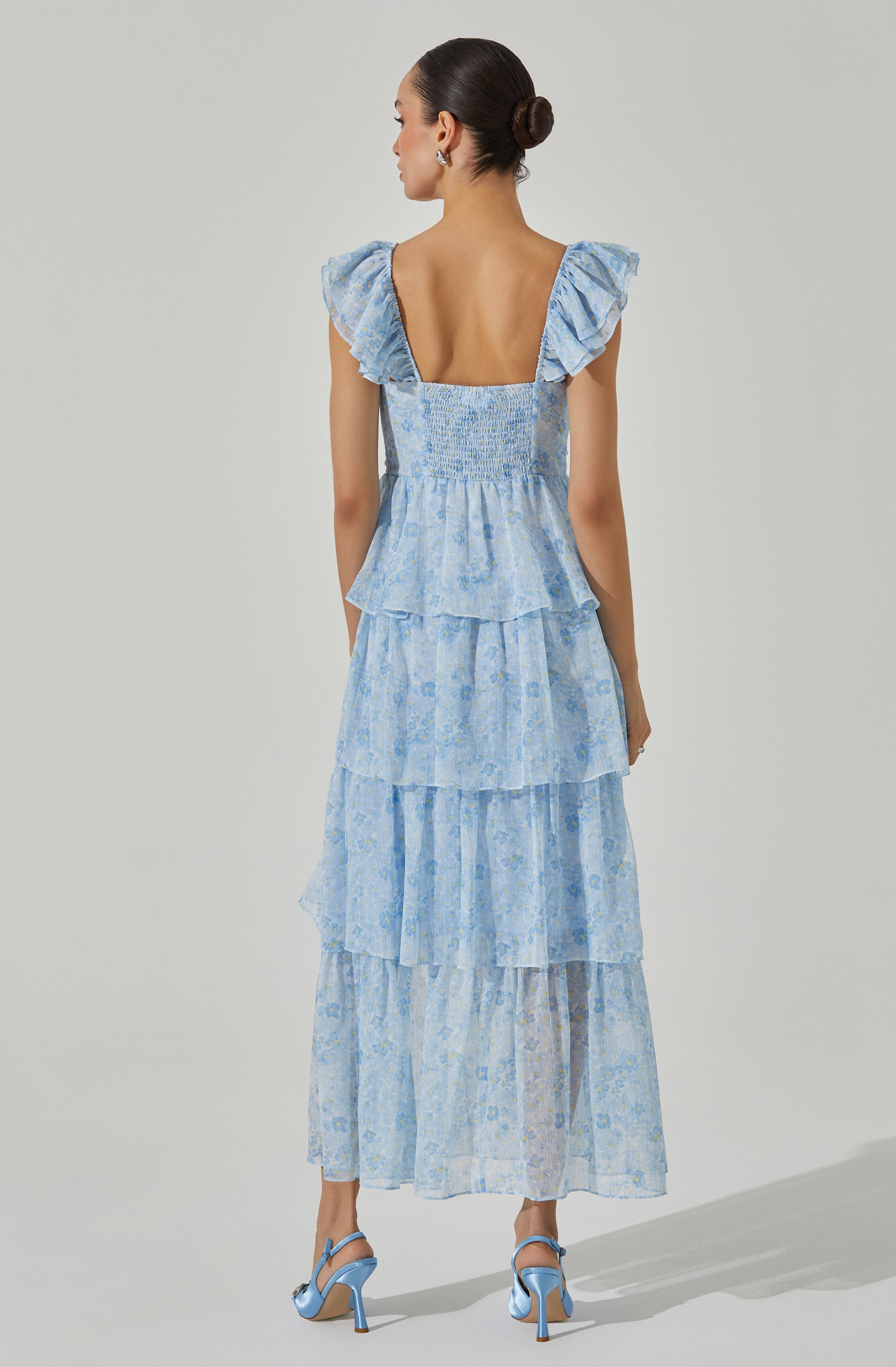 A woman is wearing a light blue, tiered dress with a floral pattern. The dress features smocked fabric at the chest, ruffled cap sleeves, and a deep square neckline at the back. She is also wearing matching light blue high-heeled sandals.