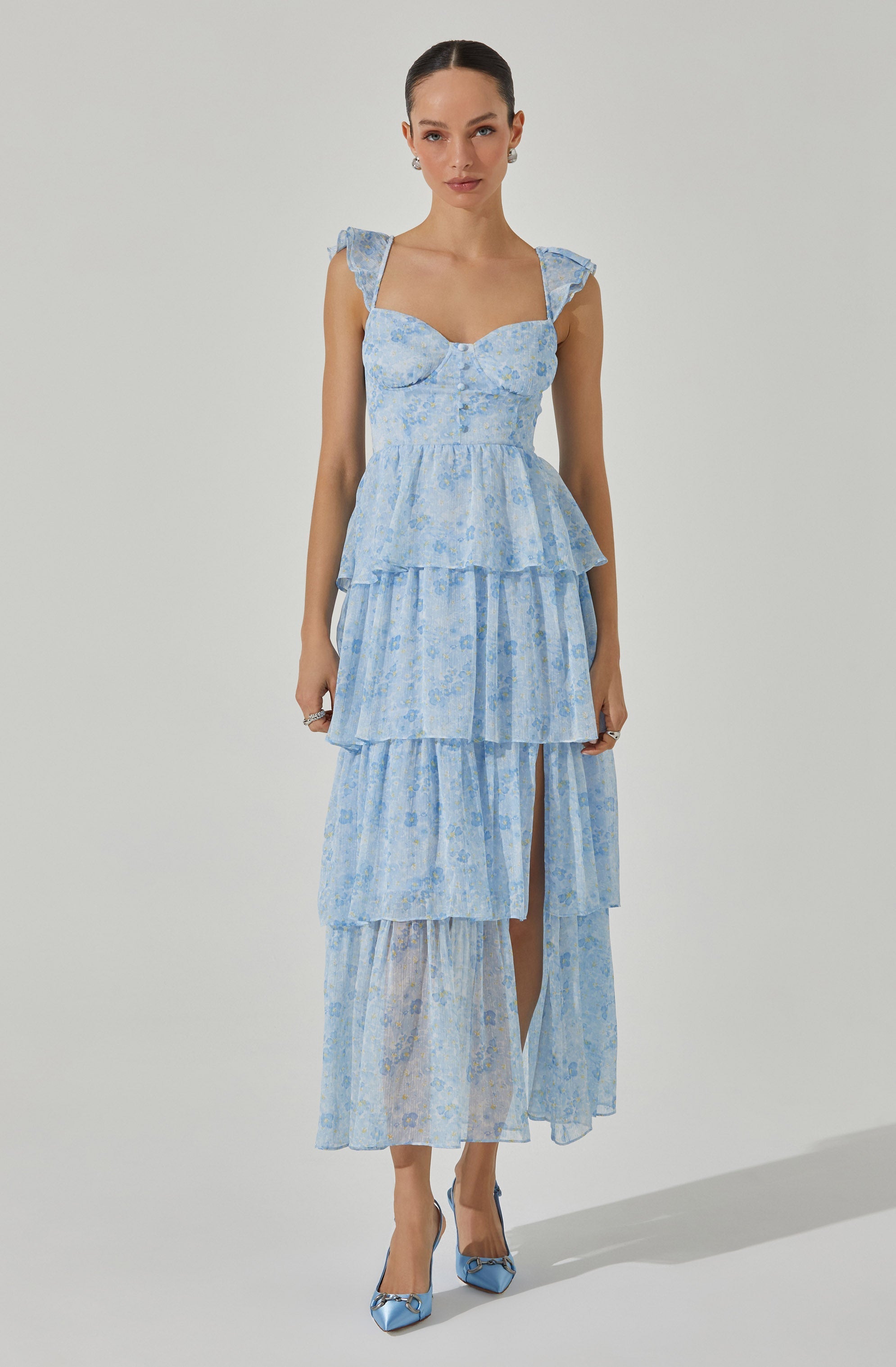 The woman is wearing a light blue, tiered maxi dress with a delicate floral pattern. The dress features ruffled shoulder straps, a fitted bodice with a sweetheart neckline, and multiple layered skirts with a slit showing her right leg. She also has on matching light blue high heels with bow details.