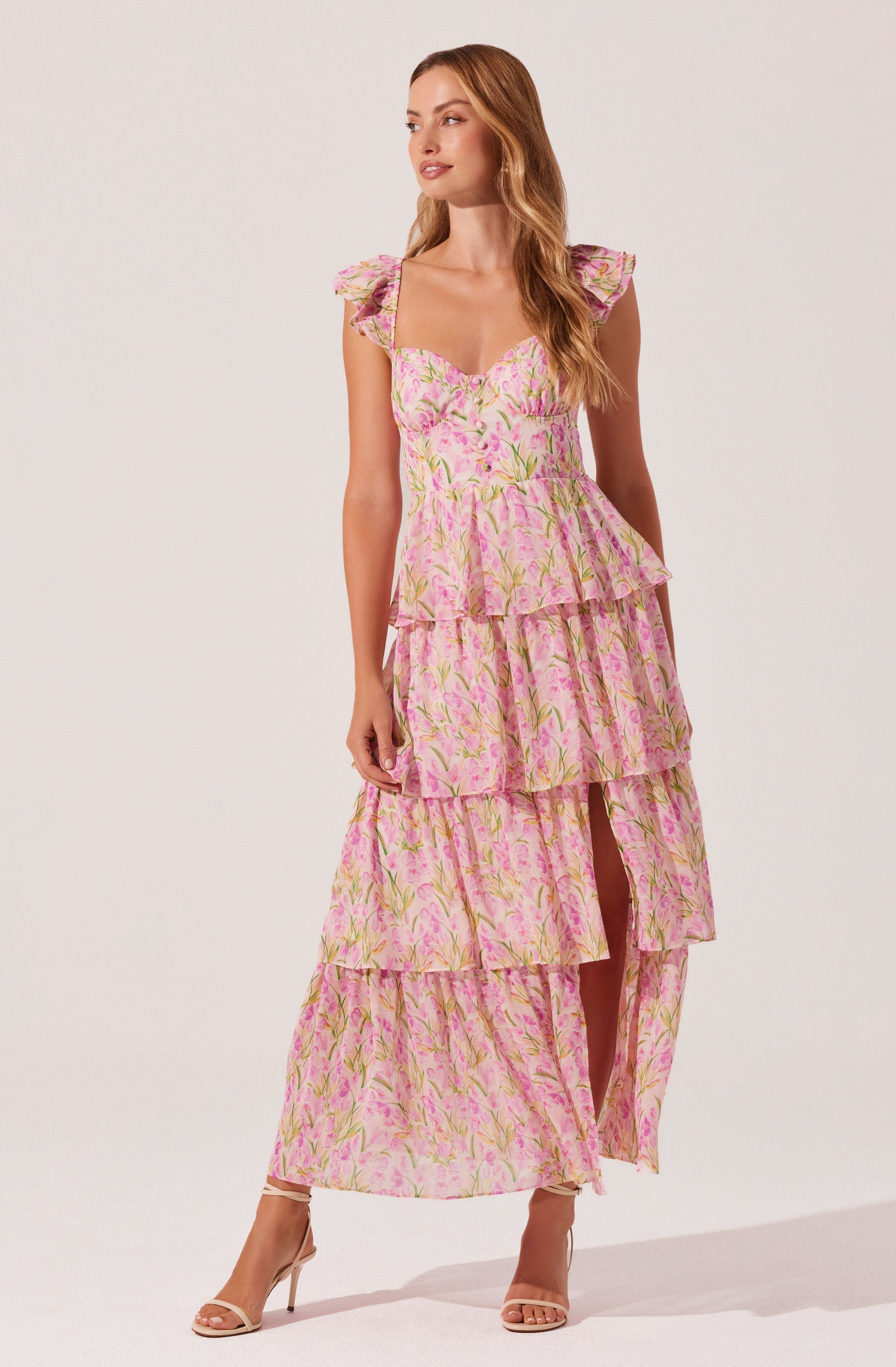 The dress is a sleeveless, full-length garment with a fitted bodice and a square neckline. It features shoulder ruffles and a tiered skirt with four layers of ruffles. The fabric has a pink background with a floral pattern in green and yellow tones. The dress has a high slit in the front, revealing part of the legs. The model is wearing nude-colored high-heeled sandals.