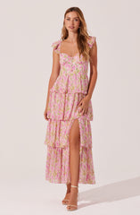 A woman is wearing a long, flowy pink dress with a floral pattern. The dress features ruffled cap sleeves, a square neckline, and a tiered skirt with a thigh-high slit on the left side. She is also wearing nude high-heeled sandals.