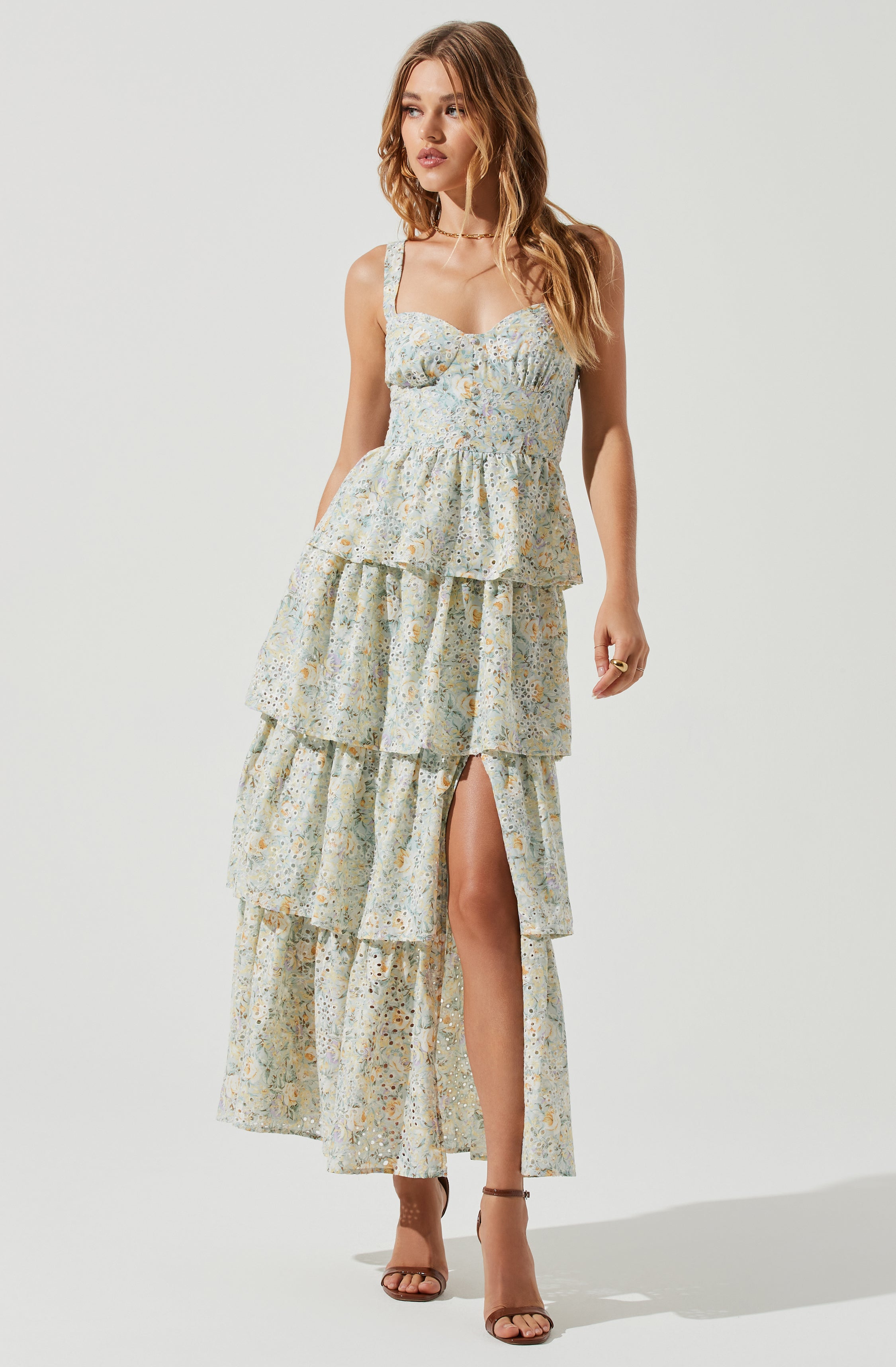 A woman is wearing a sleeveless, floral-patterned maxi dress with multiple tiered ruffles. The dress has a fitted bust area, a high waistline, and a slit on one side revealing her leg. She is also wearing open-toe high-heeled sandals.