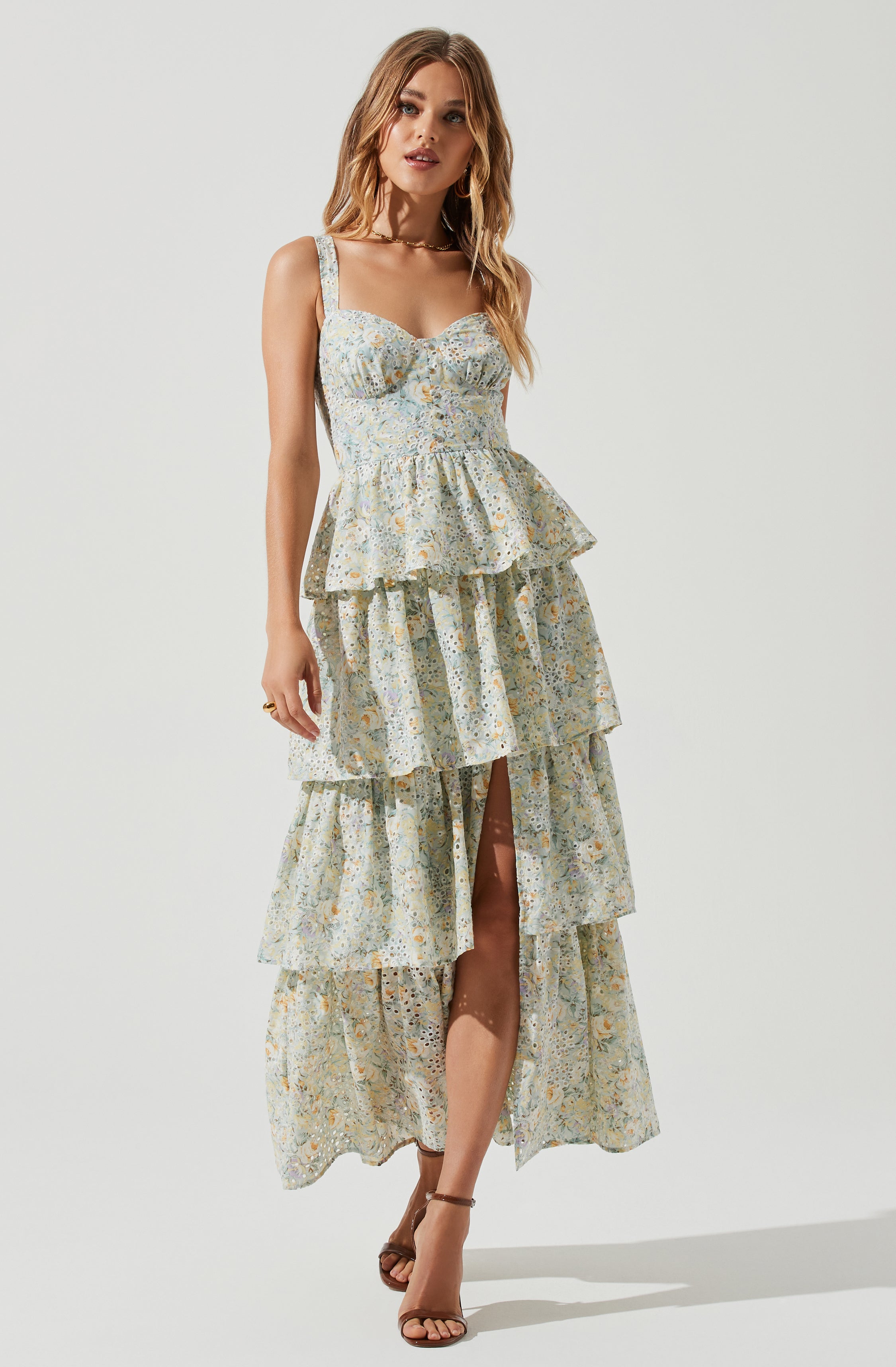 A woman is wearing a sleeveless, multi-tiered dress with puffed bodice and square neckline. The dress features a light-colored, intricate floral and abstract pattern throughout. She pairs the dress with open-toed, strappy high heels.