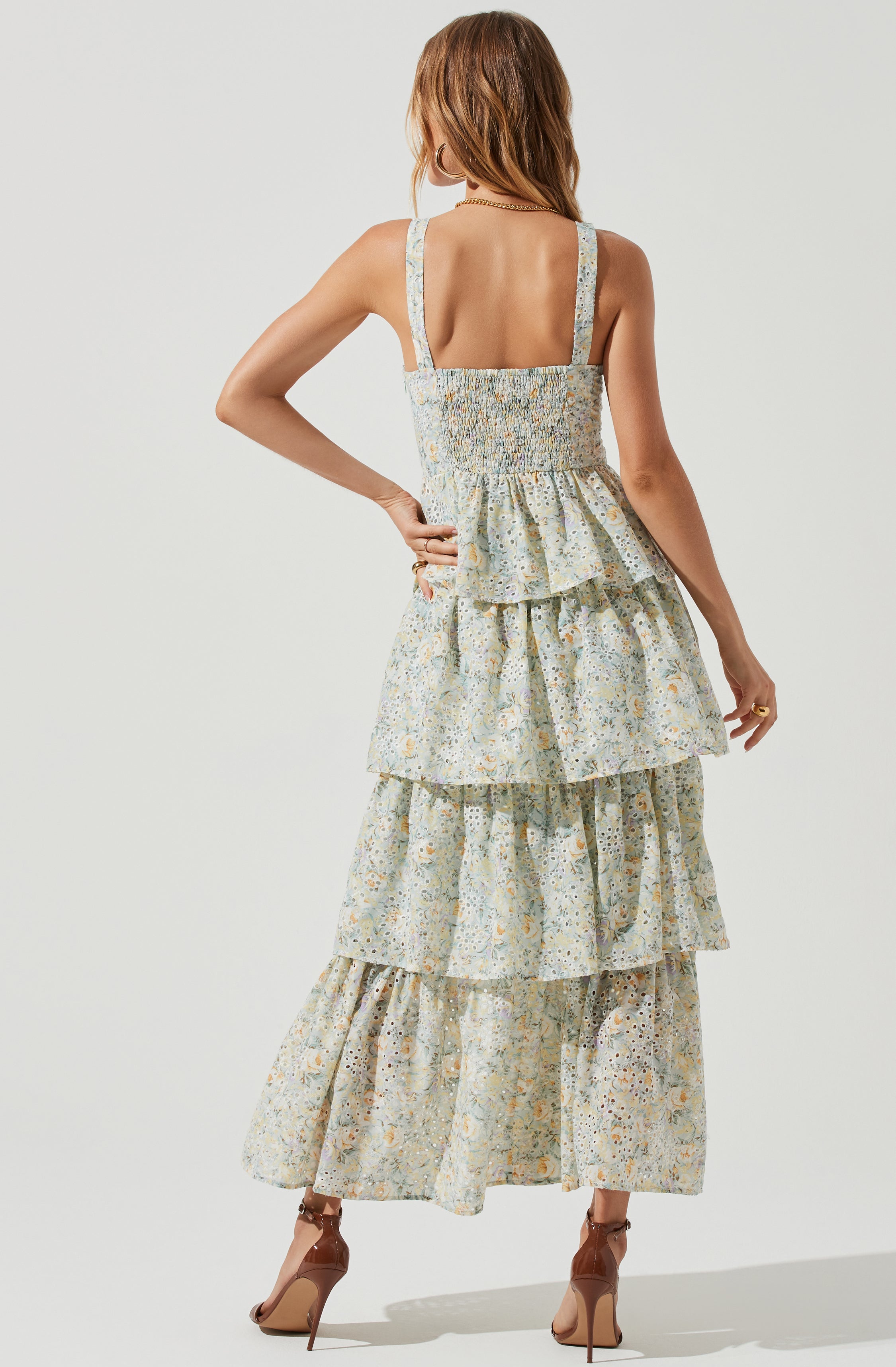 The dress is a sleeveless, layered maxi with wide shoulder straps, featuring a light, floral, and patterned fabric in soft pastel shades. The top portion of the dress has a gathered, smocked bodice, and the skirt sections are composed of multiple tiered ruffles, adding volume and movement. The overall look is feminine and summery. The model is also wearing nude high heels.