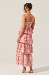 A woman is wearing a sleeveless, floral maxi dress with multiple ruffled layers. The dress features thin shoulder straps and a fitted bodice, with a pattern of pink, red, and orange flowers on a light background. She is also wearing a pair of white heeled shoes and gold earrings. Her hair is styled in a neat bun.