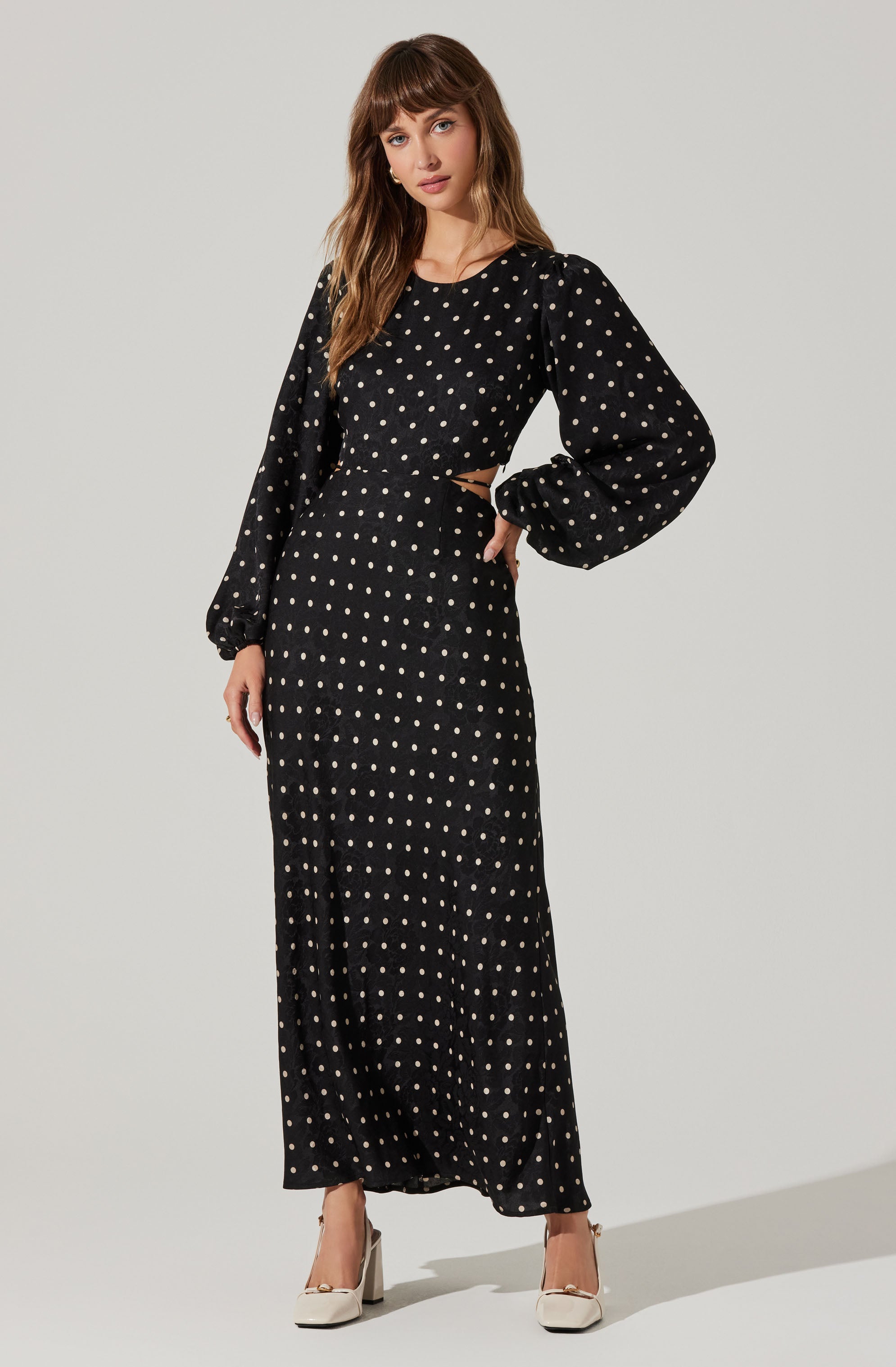 A woman wearing a black, long-sleeved maxi dress with a white polka dot pattern, featuring puffed sleeves that gather at the wrists, and a fitted waist. She is also wearing white high-heeled shoes.