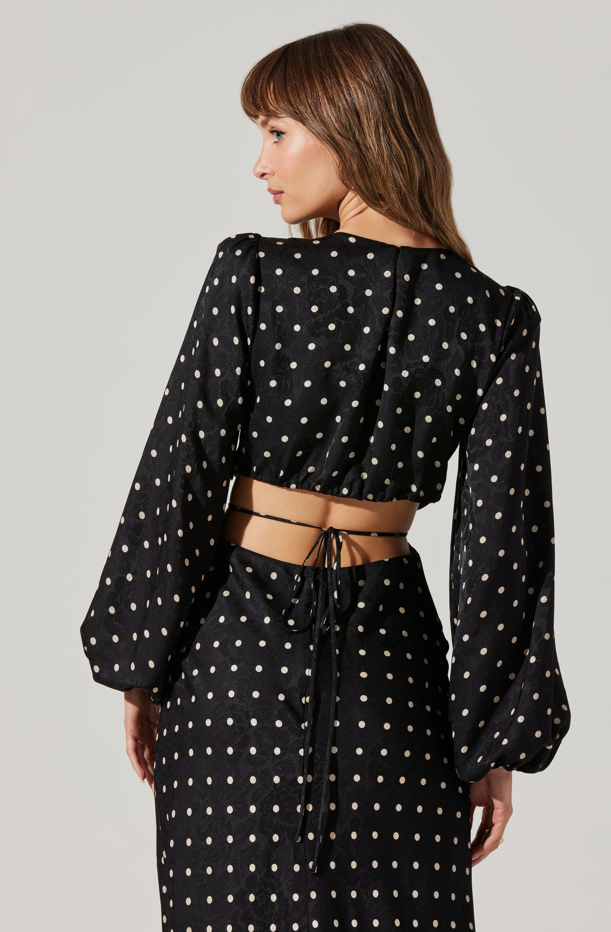 A woman is wearing a black two-piece outfit with white polka dots. The top has long puffed sleeves and a cropped length, tied at the back with a thin black string. The matching skirt has the same polka dot pattern and high waist.