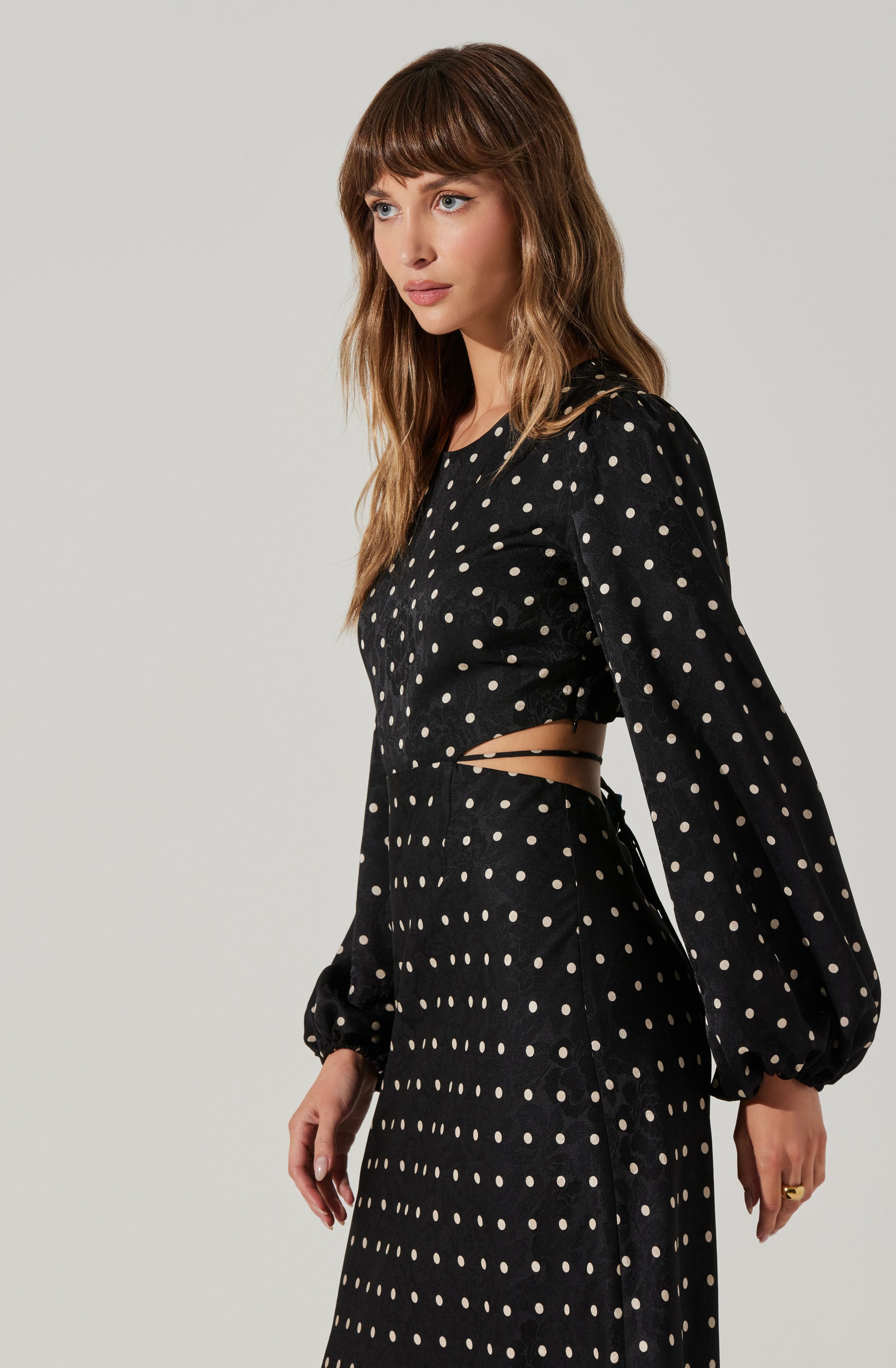 A woman is wearing a black dress with white polka dots. The dress features long, voluminous sleeves gathered at the wrists, a cutout on the side revealing her waist, and a high-waisted skirt. The overall style is elegant with a vintage-inspired design.