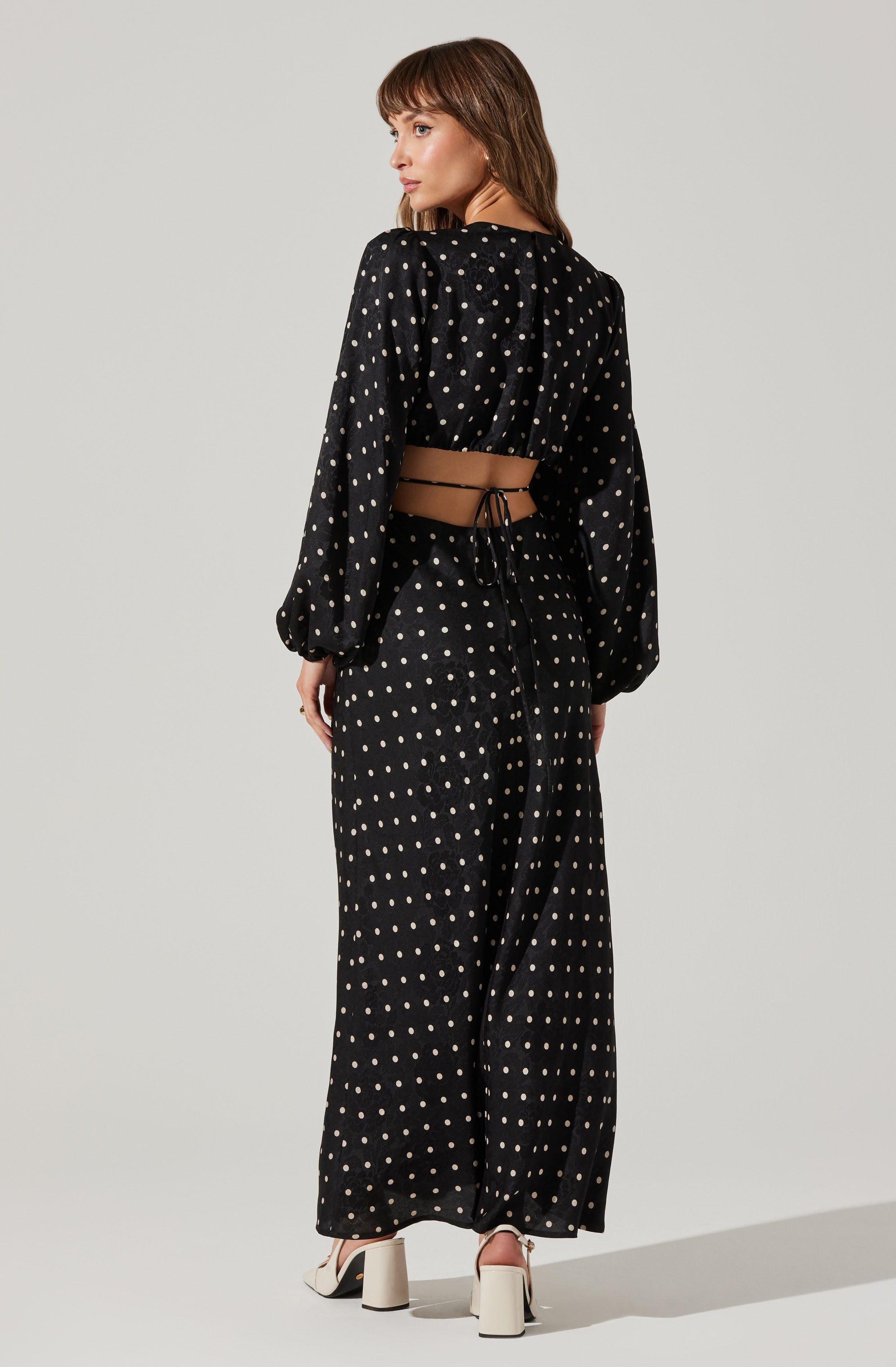 A woman is wearing a two-piece black outfit with white polka dots. The top has long, puffed sleeves and a drawstring at the back, leaving her midriff exposed. The matching skirt is long and flowing, reaching her ankles. She is also wearing white high-heeled shoes with chunky heels.