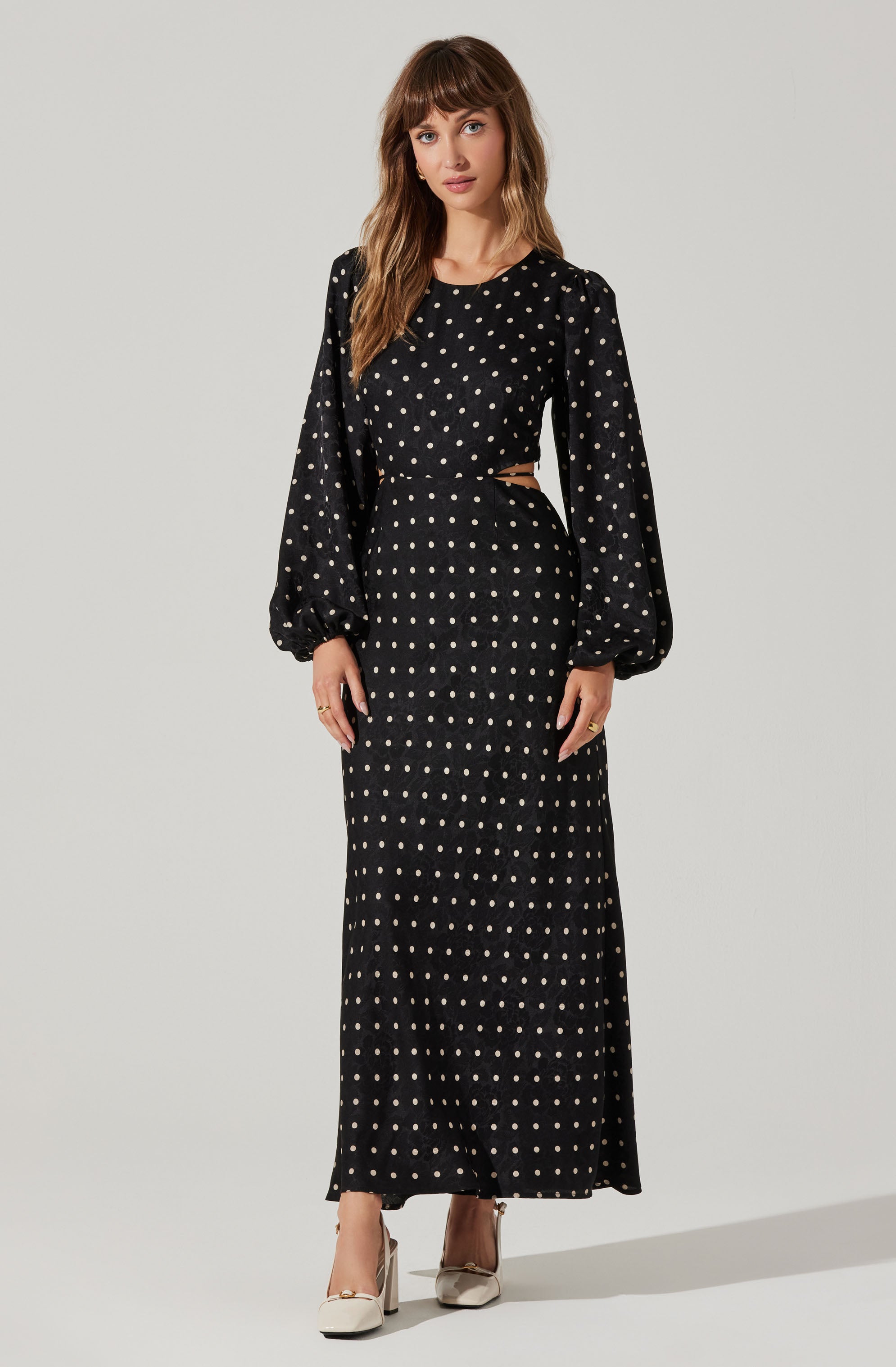 A woman is wearing a black dress with white polka dots. The dress features long, puffy sleeves gathered at the wrists, a fitted waist, and a cut-out detail at the waistline. She is also wearing white high-heeled shoes with a bow detail.