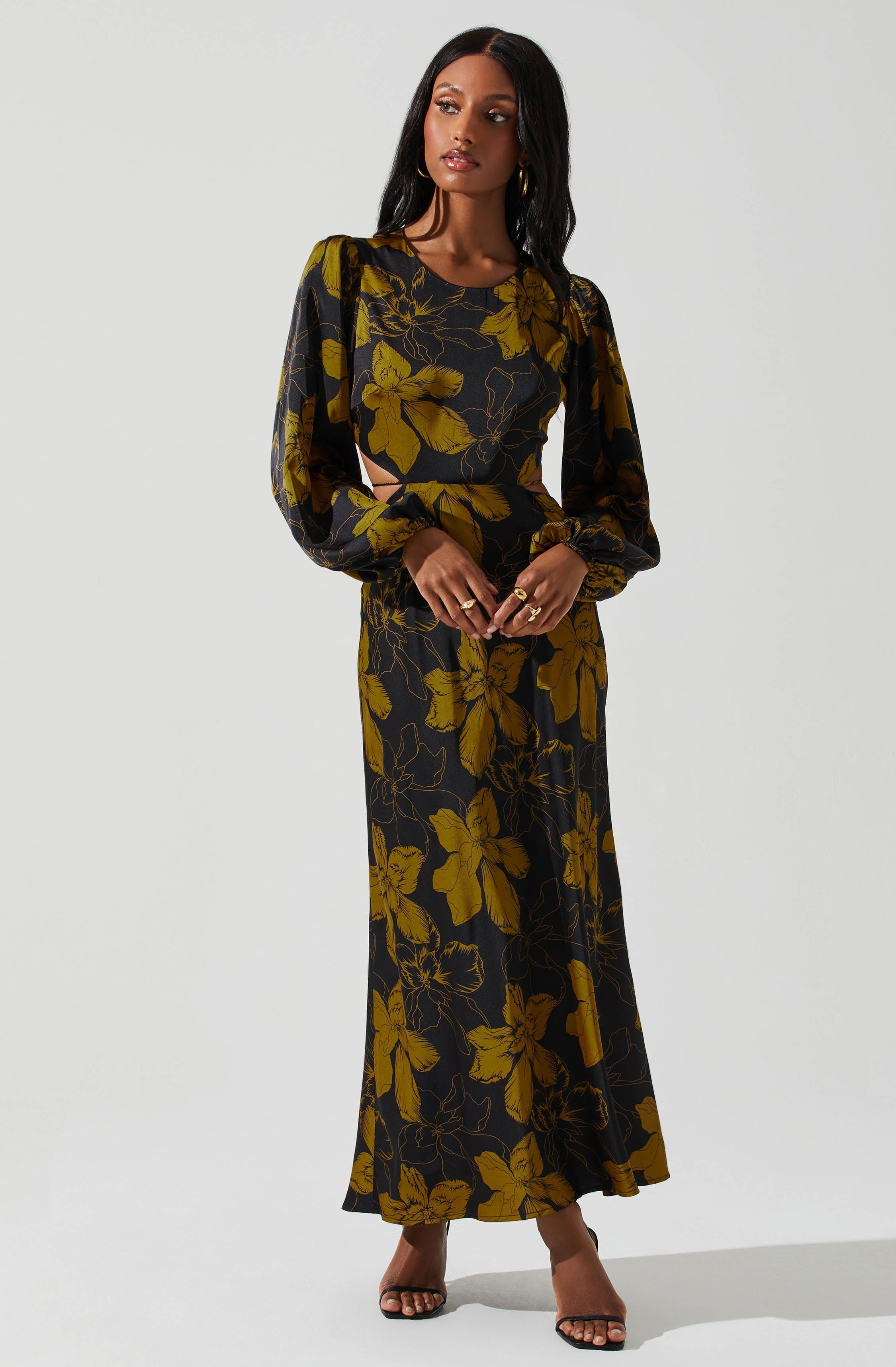 A woman is wearing a long black dress with a large yellow floral pattern. The dress has long, puffed sleeves that gather at the wrists, a round neckline, and a cutout at the waist on one side. She is accessorized with gold hoop earrings and rings, and she is wearing open-toed black heels.