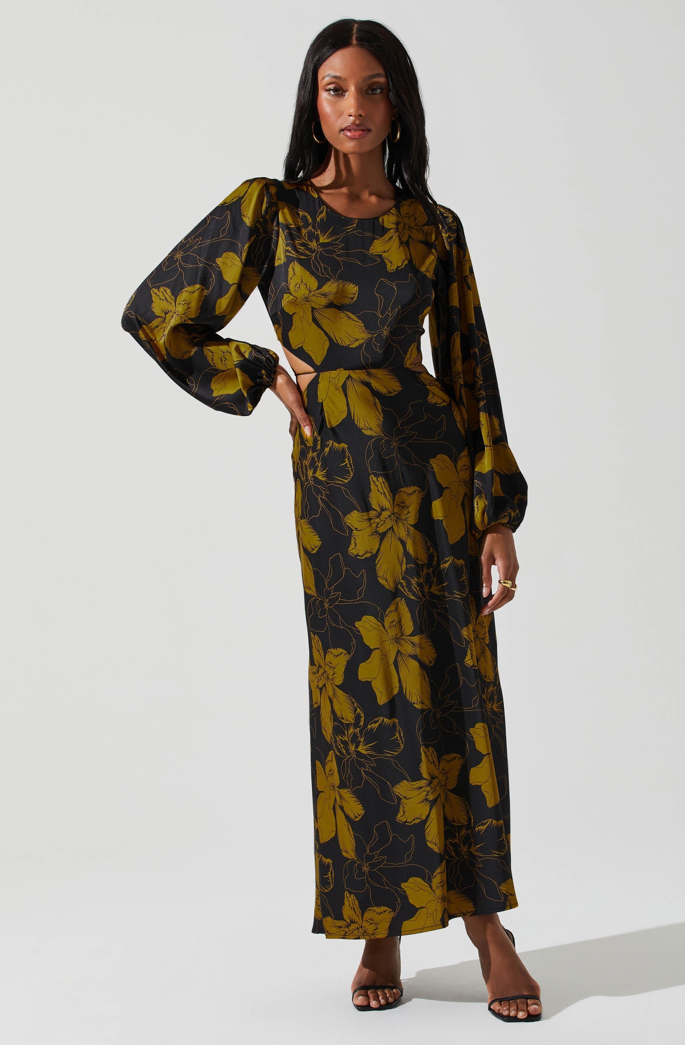 A woman is wearing a long black dress with large yellow floral patterns. The dress has long, puffed sleeves with elastic cuffs and a fitted bodice. She pairs the outfit with black open-toe heels and gold hoop earrings.