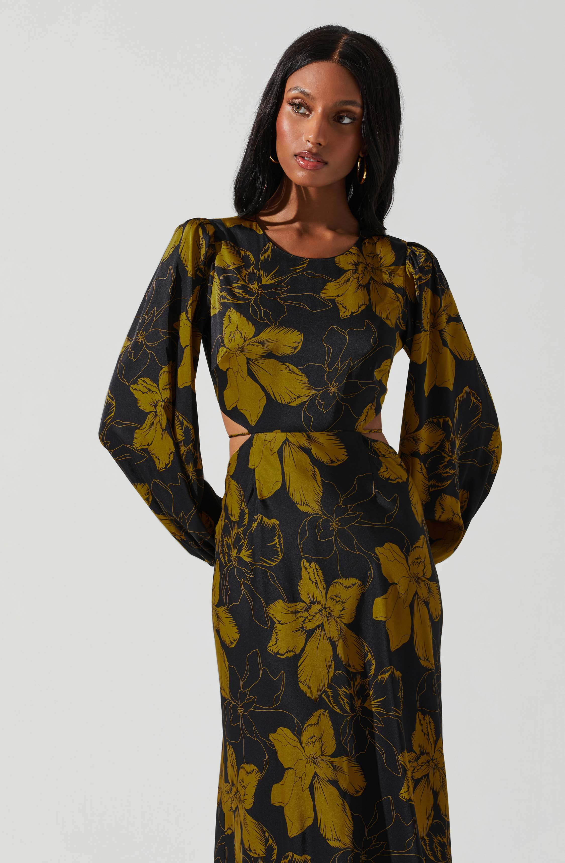 A woman wearing a long black dress with large, mustard-yellow floral patterns. The dress features puffed long sleeves, a fitted waist, and cut-out sections at the sides.