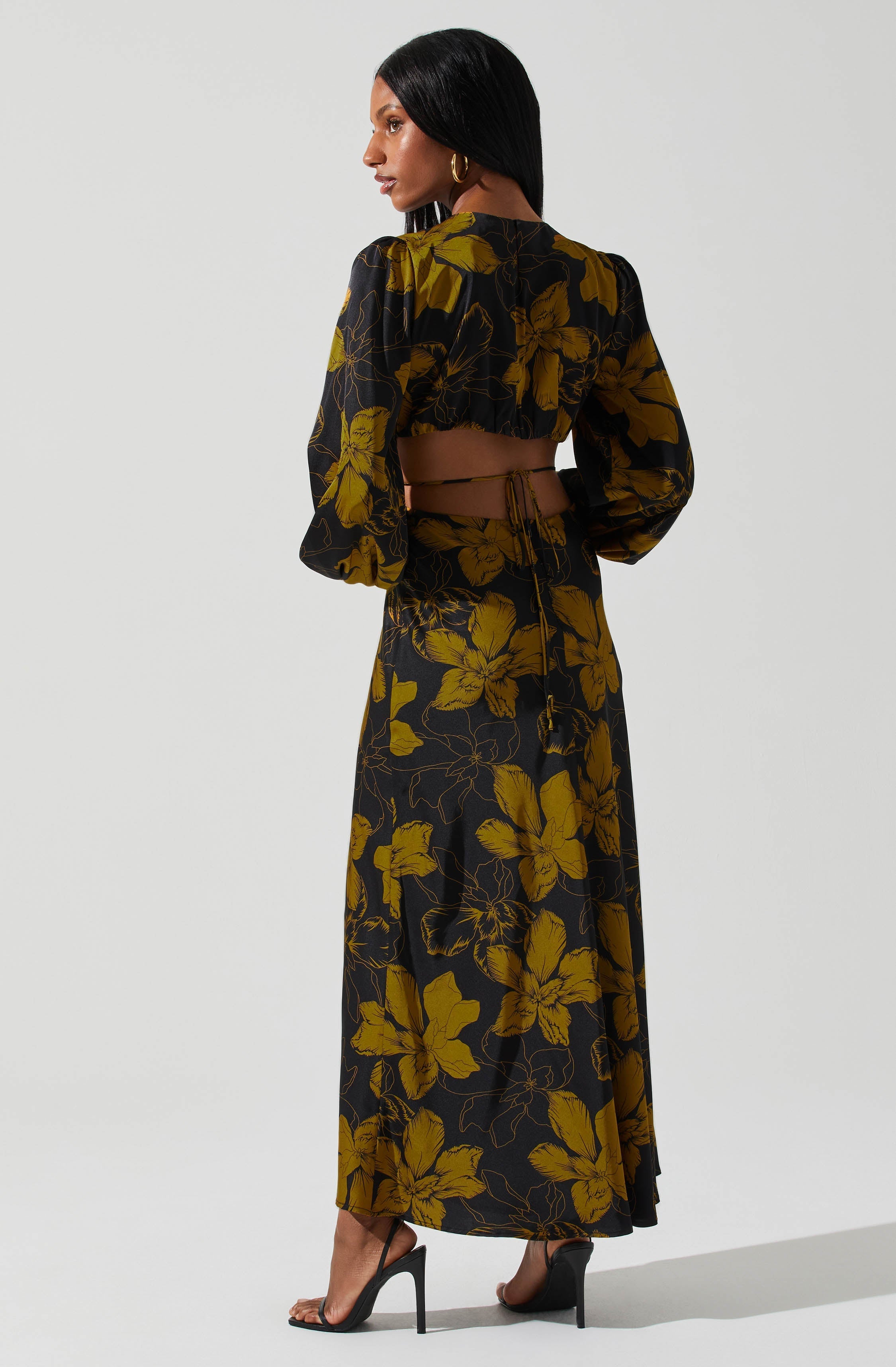 A woman is wearing a two-piece outfit with a black background and large yellow floral patterns. The top has long, puffed sleeves and a cut-out detail at the back, secured with a tie. The matching long skirt features the same floral print and reaches down to her ankles. She is also wearing black high-heeled sandals.