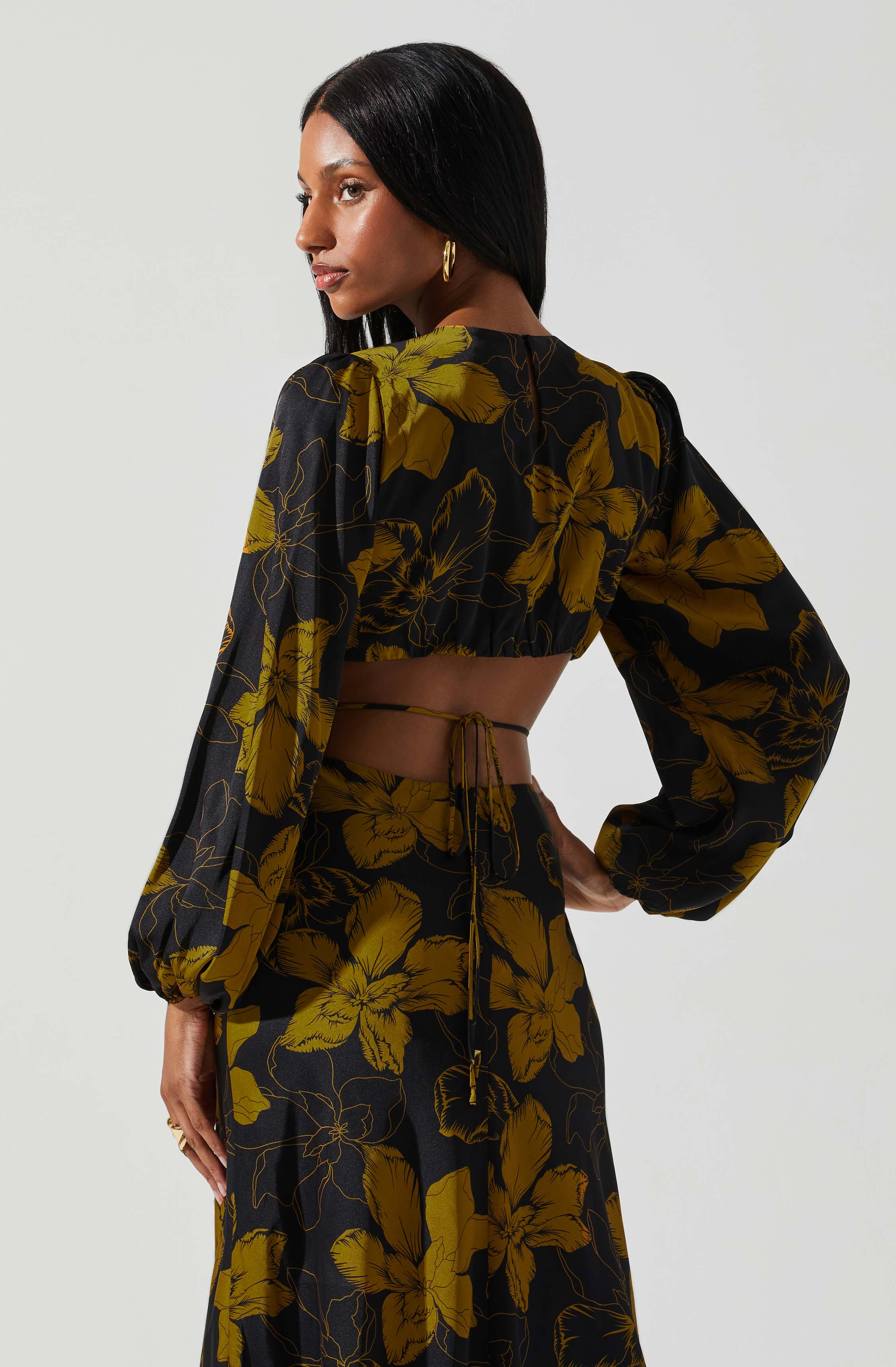 A woman with long, straight black hair wearing a black and yellow floral two-piece outfit. The top features long, puffed sleeves and is cropped, exposing a small section of her torso. The bottom is a matching high-waisted skirt with the same yellow floral pattern on a black background. She accessorizes with gold hoop earrings and a ring, standing against a plain light background.