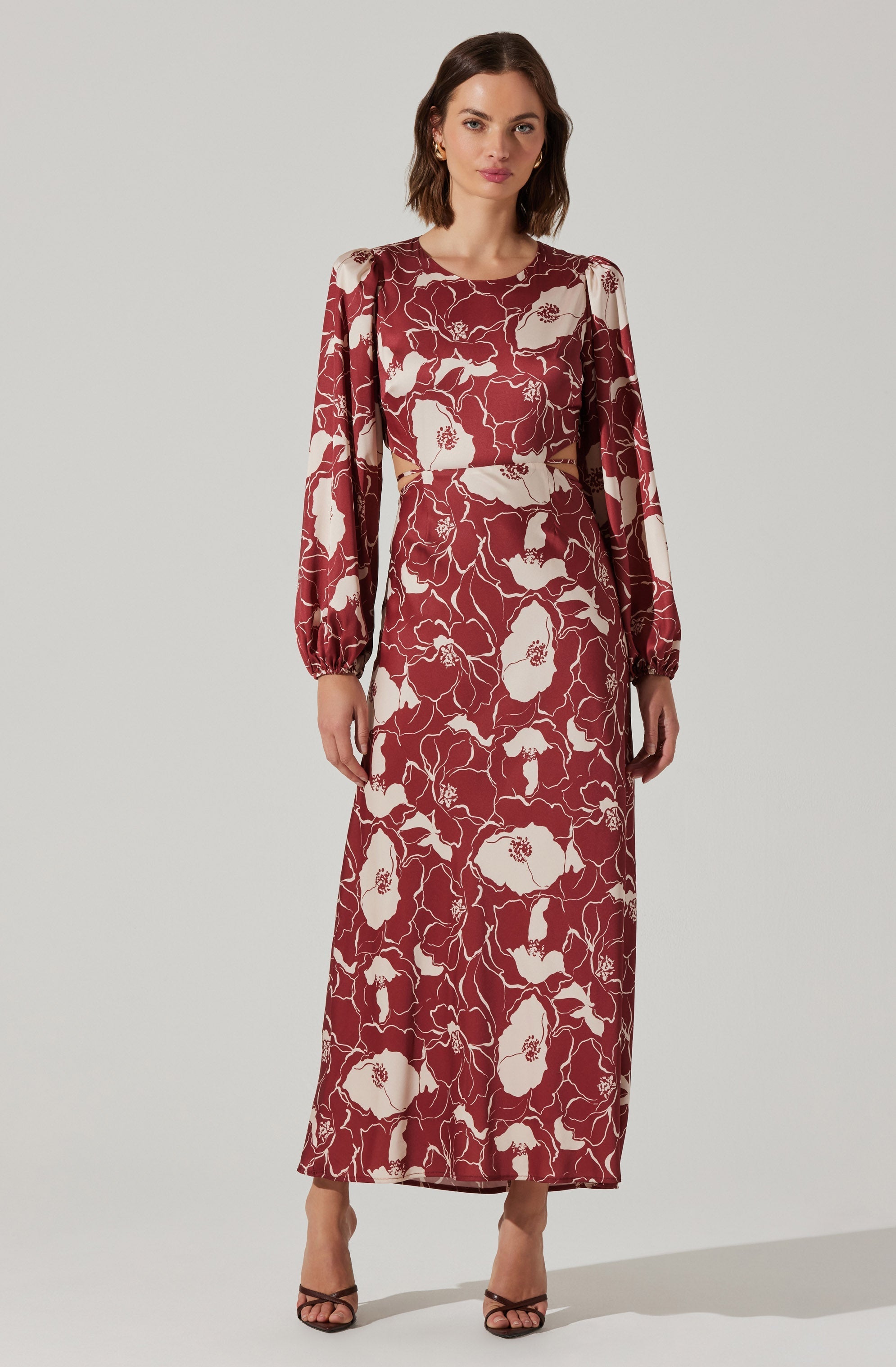 A woman is wearing a long dress with a floral pattern featuring large cream-colored flowers on a deep red background. The dress has long, puffed sleeves gathered at the wrists, a round neckline, and cut-out details at the waist. She is also wearing open-toe high heels.