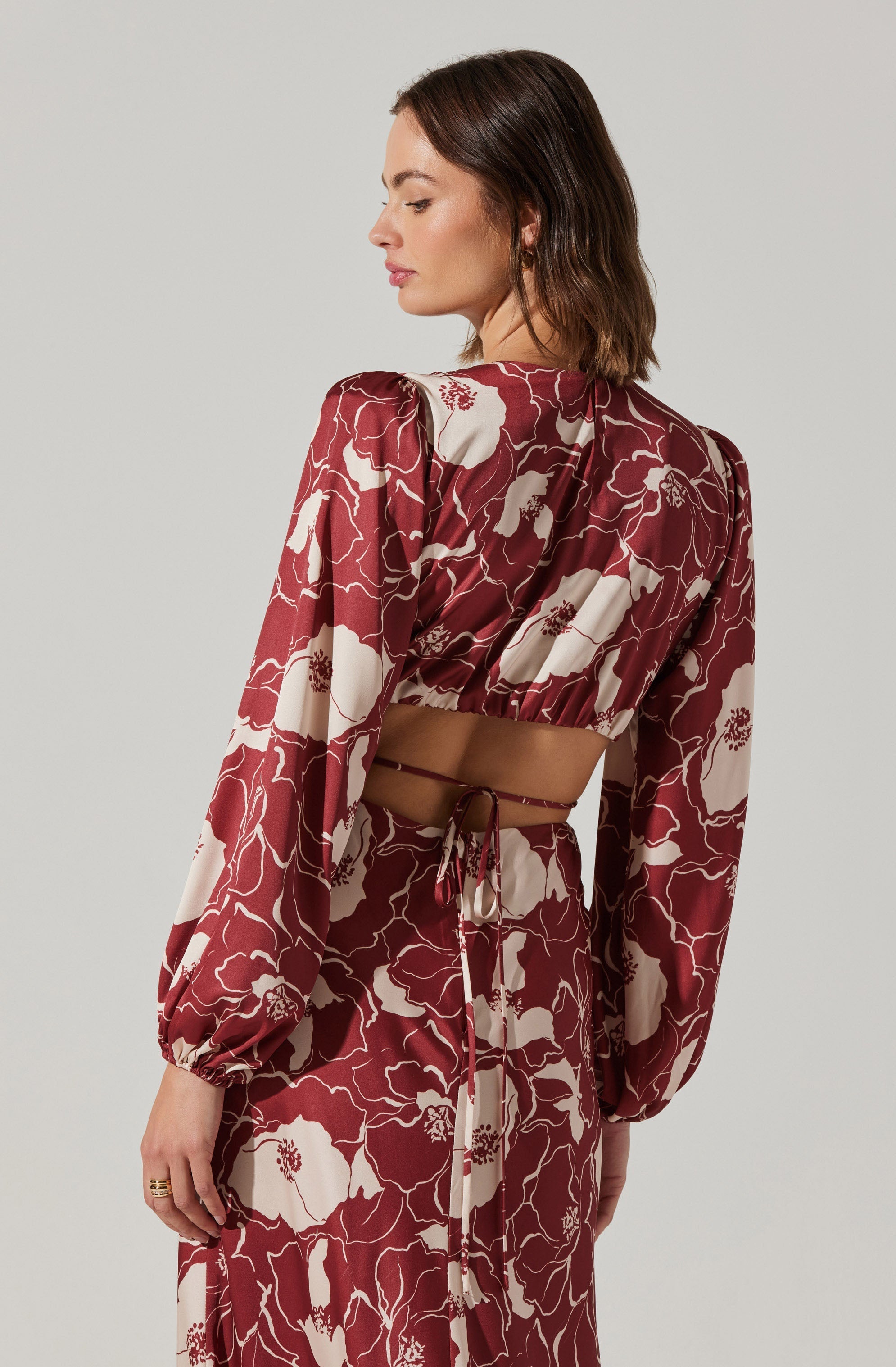 A woman is wearing a matching two-piece outfit with a large floral pattern. The top has long, voluminous sleeves that are gathered at the wrist and a cropped length, tied at the back with a thin ribbon. The skirt is high-waisted, flowing, and features the same floral print. The fabric appears lightweight and silky.