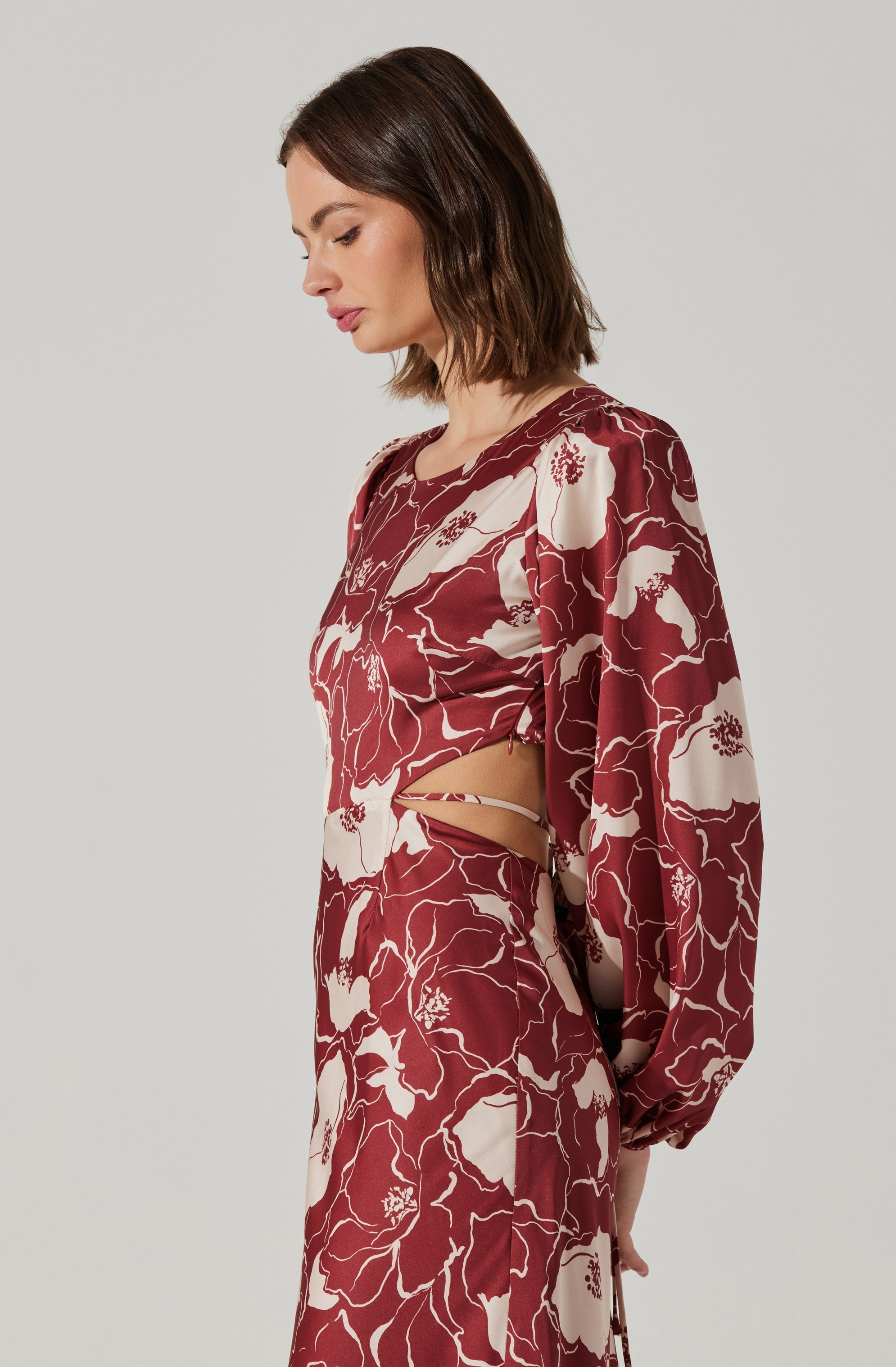 A woman is wearing a matching outfit with a deep red and cream floral pattern. The top has long, billowy sleeves and a cut-out detail on the side, revealing a hint of skin. The matching skirt is high-waisted, flowing, and features the same floral motif throughout.