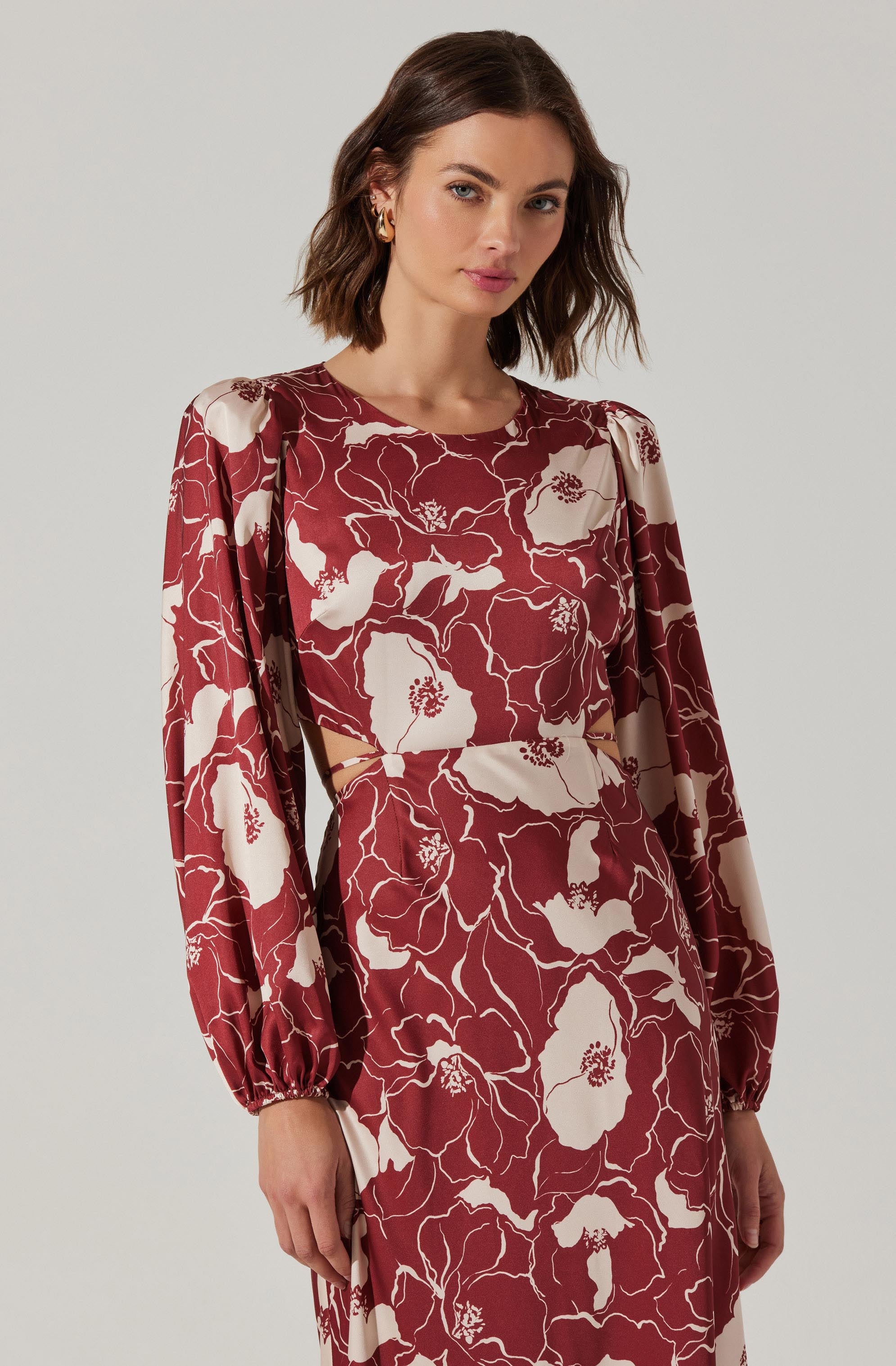 A woman wearing a long-sleeved dress with a floral pattern in shades of burgundy and cream. The dress features puffed shoulders, gathered at the wrists, and has a cut-out detail at the waist.