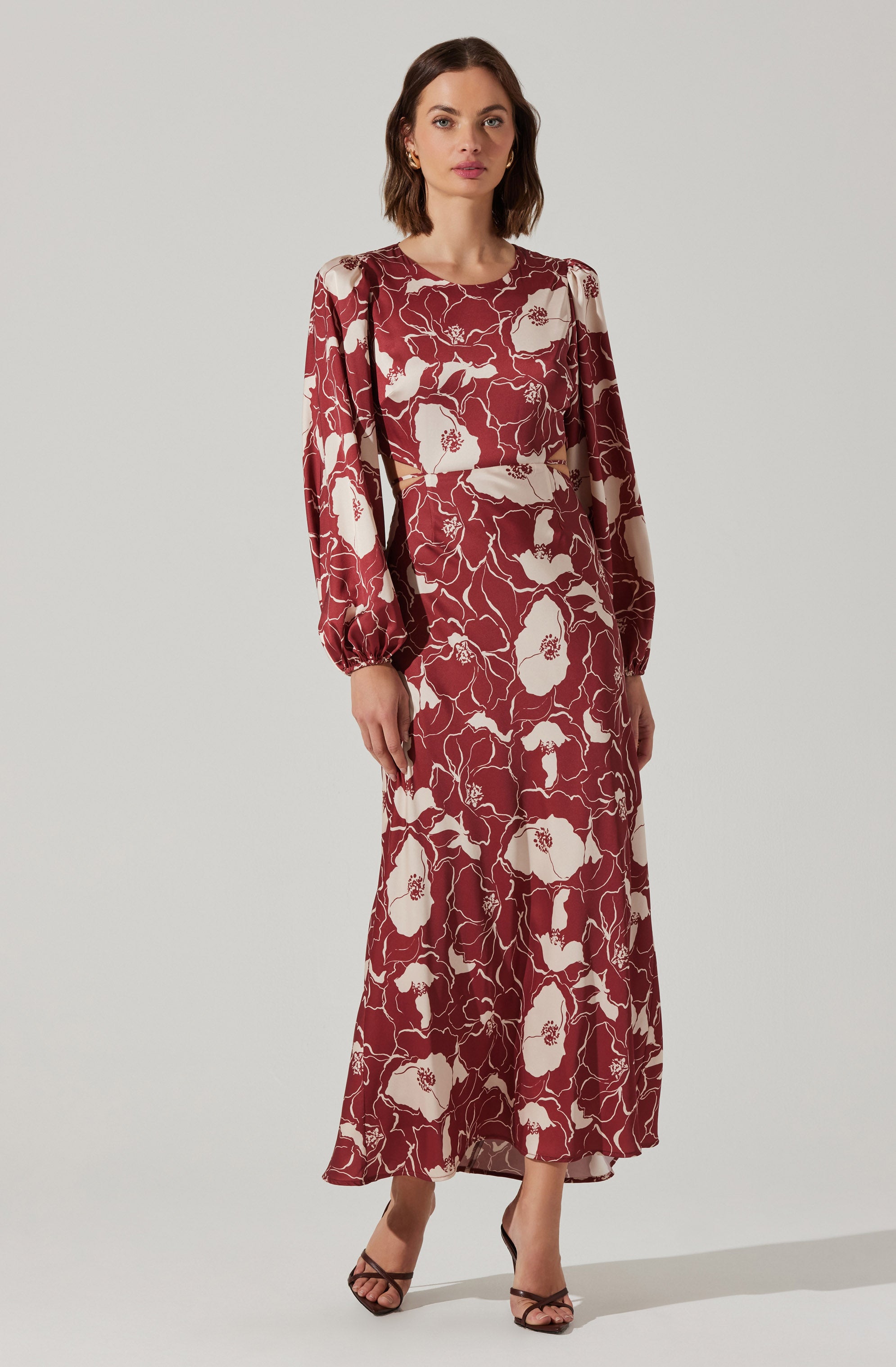 A woman is wearing a long, flowy dress with a floral pattern. The dress features a mix of cream and deep red colors, with the floral design prominently displayed throughout. The dress has long, slightly puffed sleeves with gathered cuffs, and cut-out sections at the waist. She is wearing open-toed, strappy high heels.