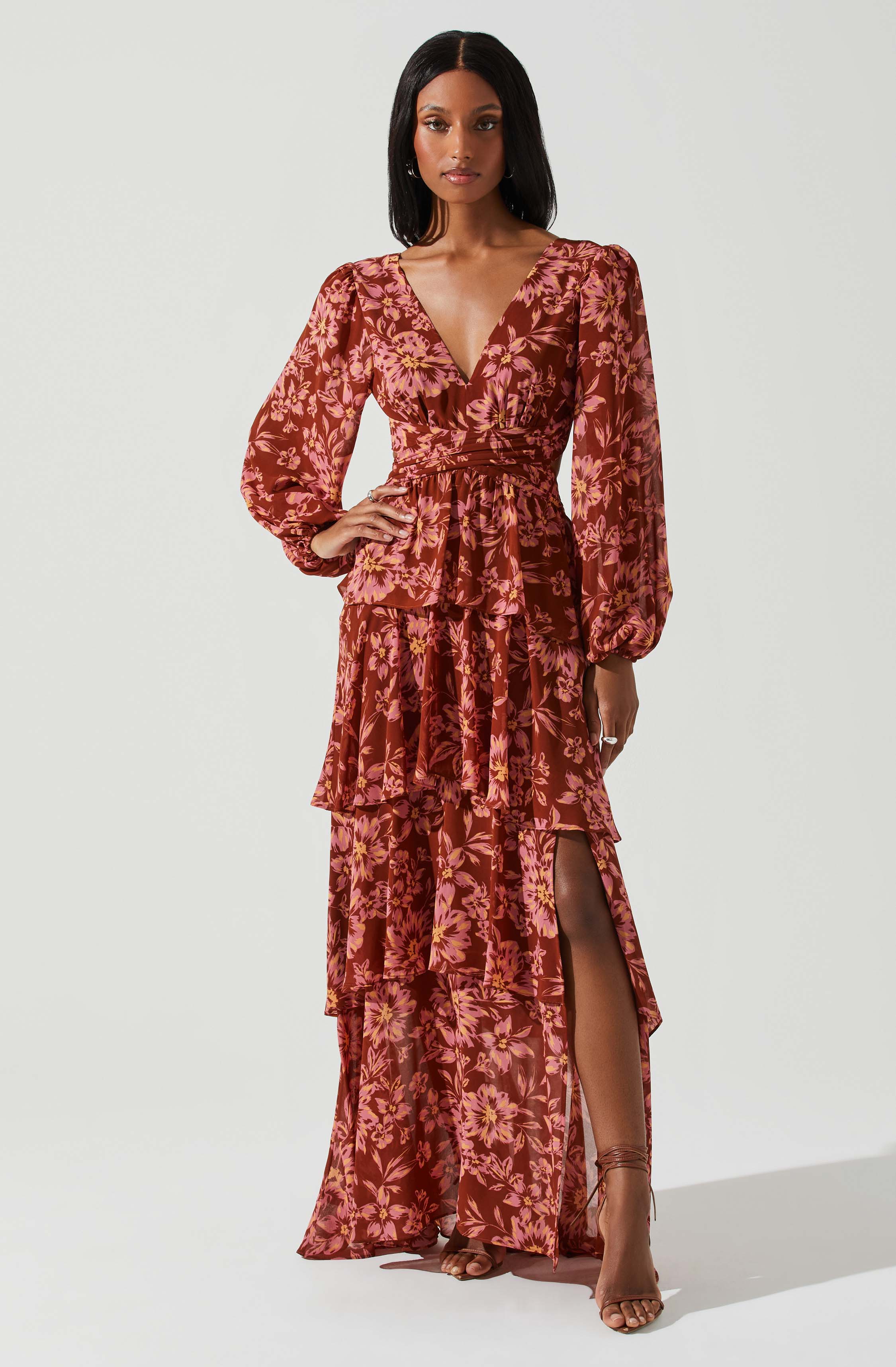 A woman is wearing a long, flowing dress with a floral pattern in shades of pink, purple, and orange on a reddish-brown background. The dress features long, puffed sleeves, a V-neckline, and a cinched waist. It has multiple tiers in the skirt, some with a high slit revealing her leg and strappy high-heeled sandals.