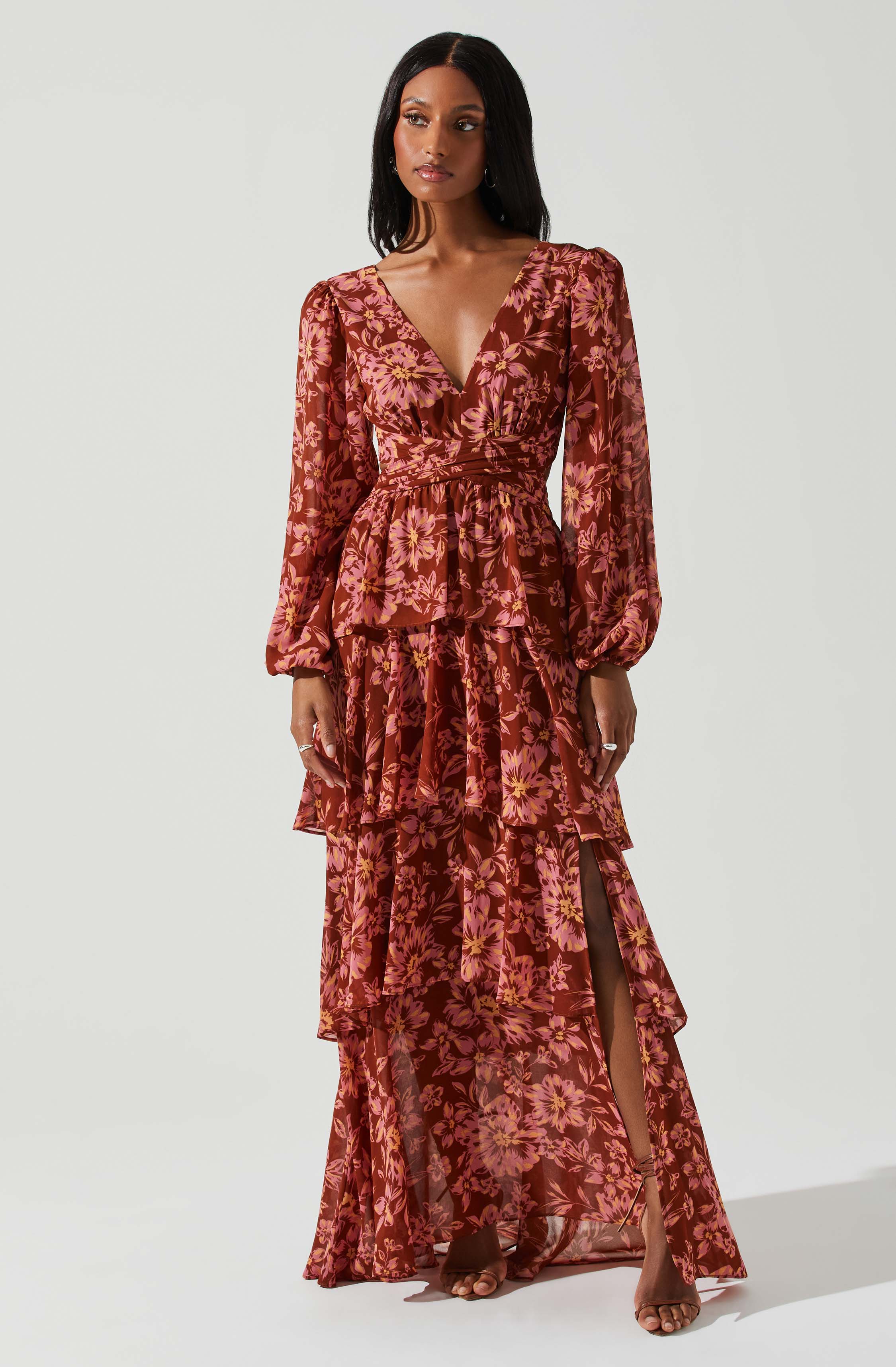 A woman is wearing a long, flowing dress with a deep V-neckline. The dress features a floral pattern in shades of pink and red, with puffed long sleeves that gather at the wrists. The waist is fitted, and the skirt has multiple tiers with ruffled edges, including a high slit on one side revealing her leg.