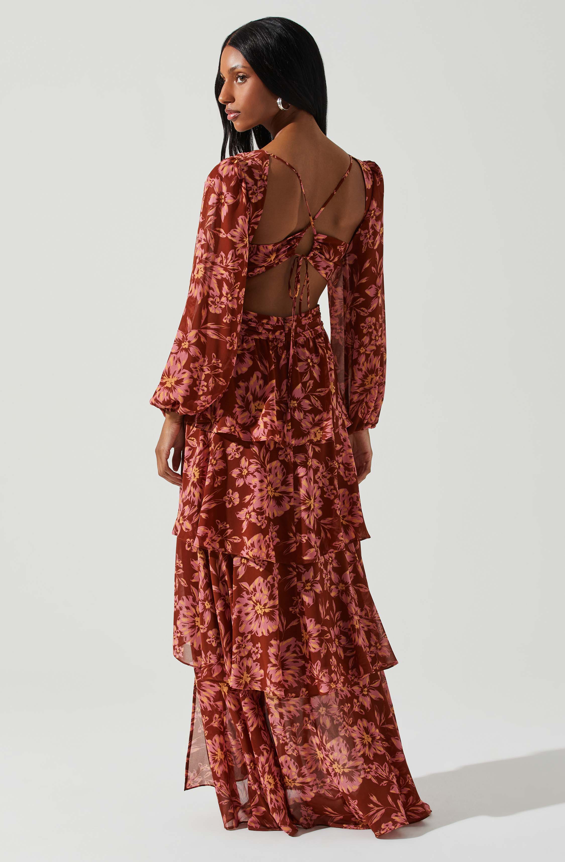 A woman is wearing a two-piece outfit with a matching top and long skirt made of sheer fabric. The outfit has a floral pattern in shades of pink, yellow, and brown. The top features long, puffed sleeves and an open back with thin, crisscrossing straps that tie into a bow. The skirt is full-length with multiple layered ruffles.