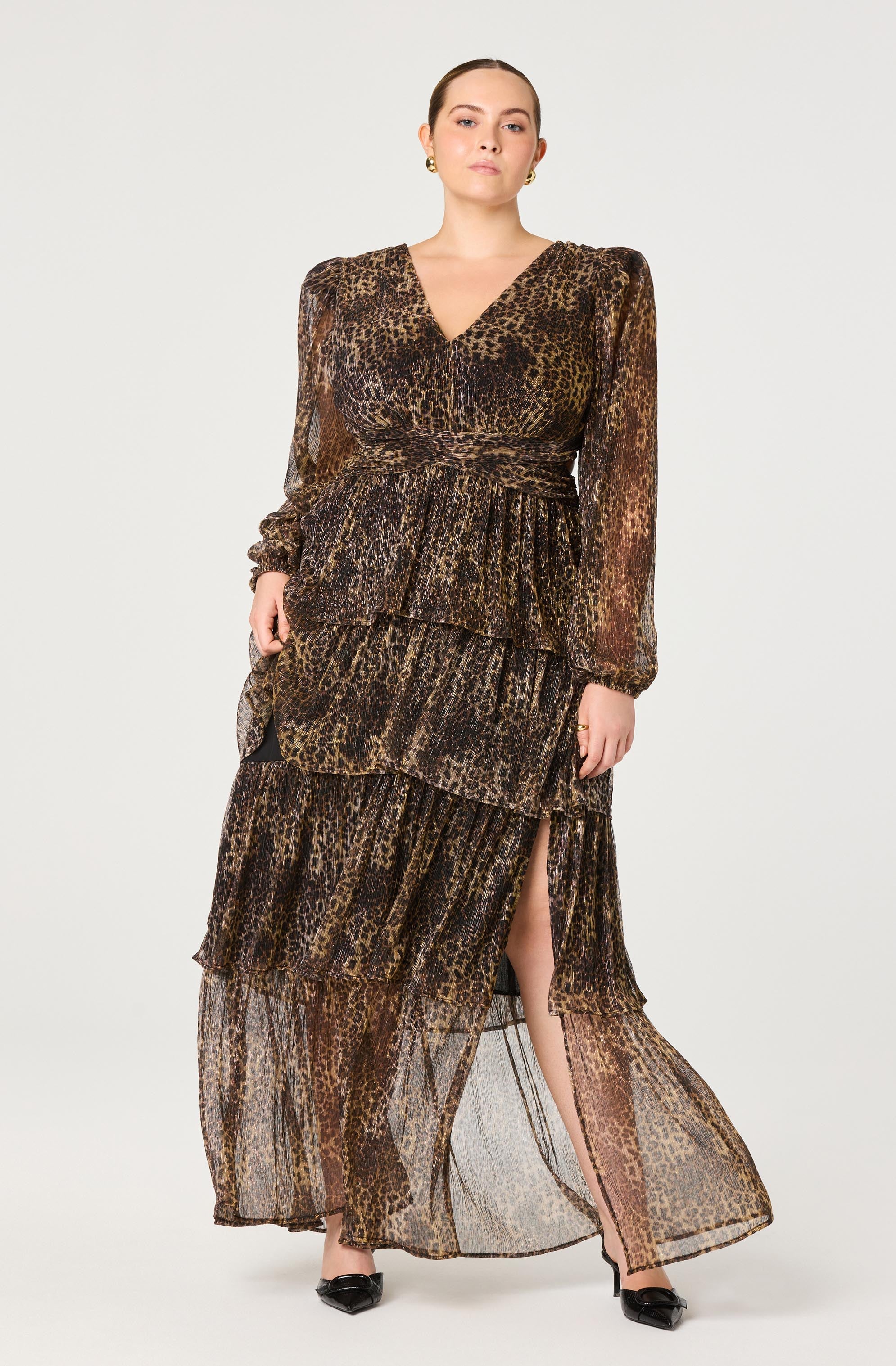 A woman is wearing a long, flowing dress with a leopard print pattern. The dress features long, sheer sleeves with gathered cuffs, a V-neckline, and multiple layered tiers with a high slit on one side, revealing her leg. The fabric is lightweight and slightly transparent.
