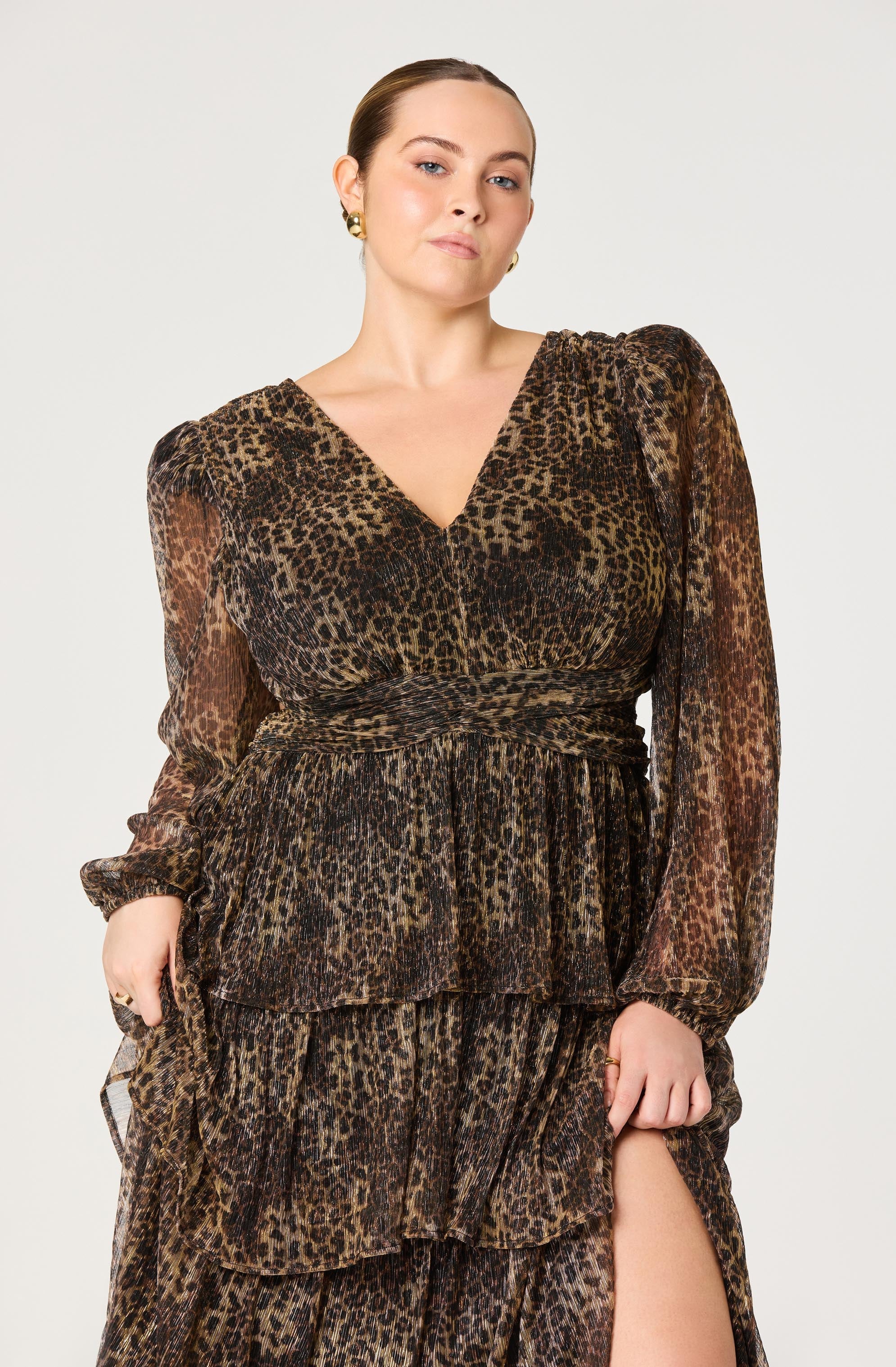 A woman is wearing a long, leopard print dress with a V-neckline. The dress features puffed shoulders, long sheer sleeves, and multiple layered ruffles on the skirt. The fabric appears lightweight and slightly sheer.