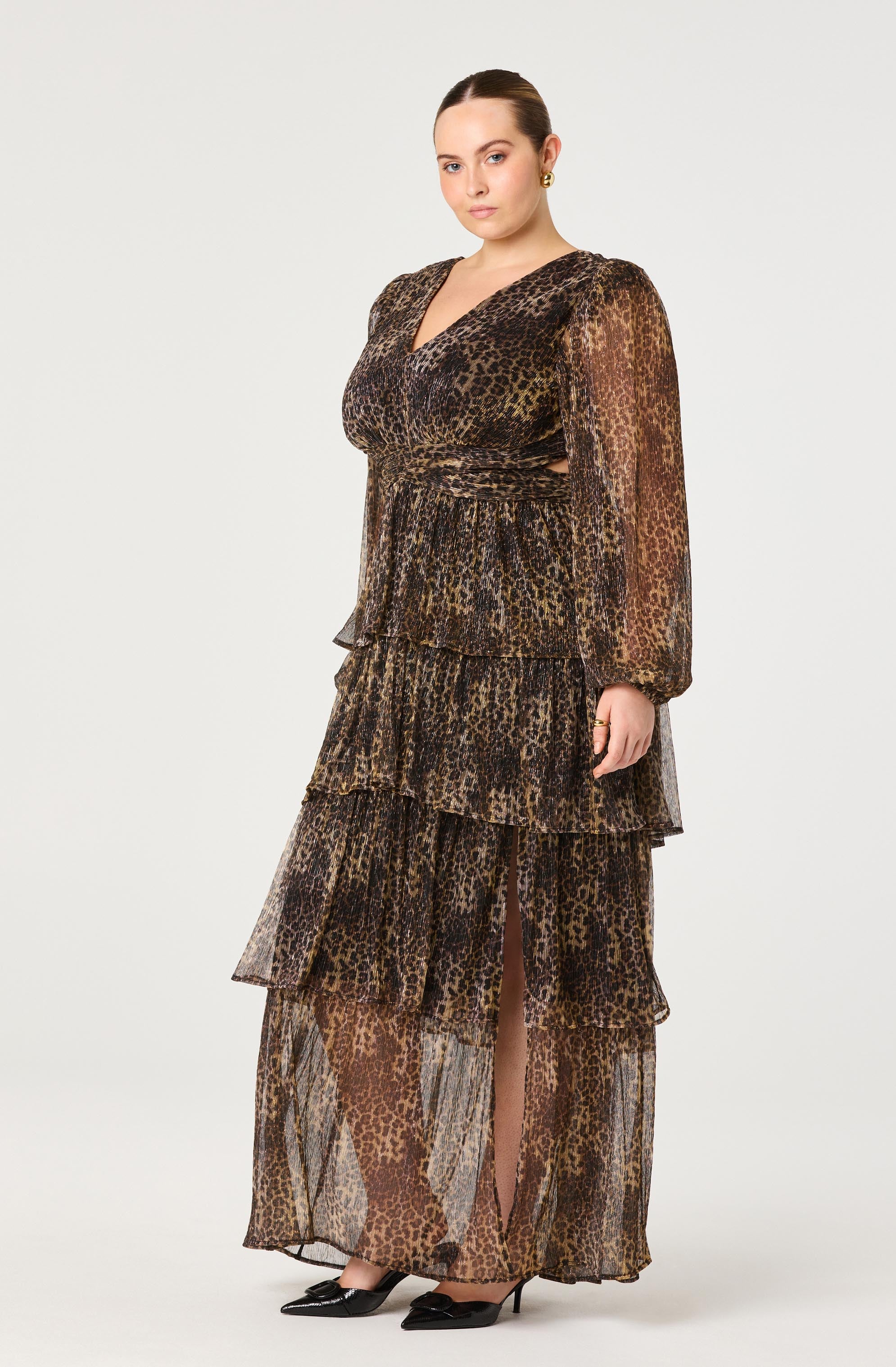 A woman is wearing a long, tiered maxi dress with a leopard print pattern. The dress has a V-neckline, long sheer sleeves, and is fitted at the waist. The fabric appears lightweight and slightly sheer, with multiple ruffled layers creating a flowing silhouette.