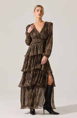 A woman is wearing a long, flowy dress with a leopard print pattern. The dress has puffed long sleeves, a V-neckline, and a fitted waist. It features multiple tiered ruffles down the length, with a high slit on one side, revealing knee-high black boots with high heels.