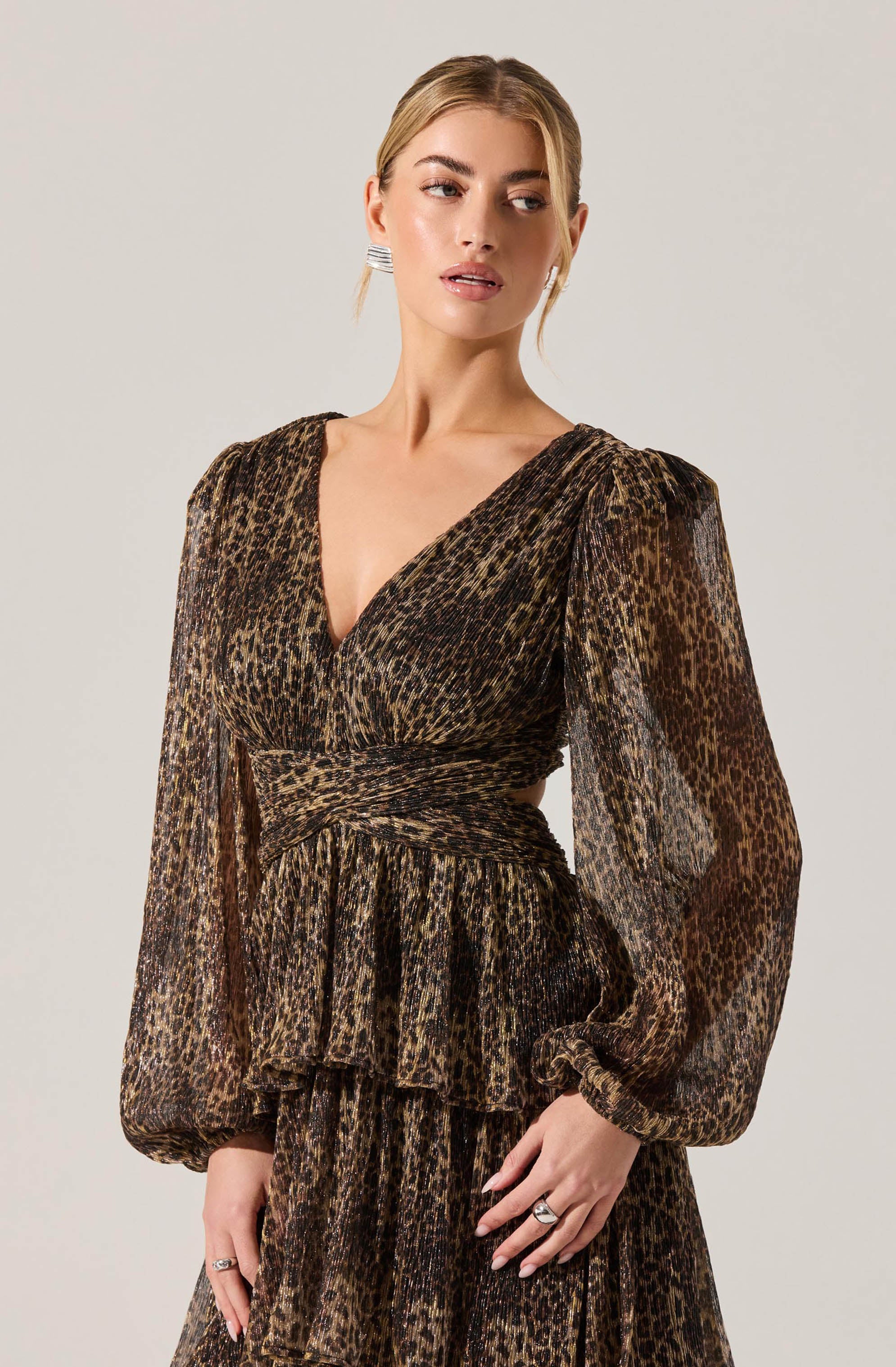 A woman is wearing a long-sleeved dress with a leopard print pattern. The dress features a deep V-neckline, puffed sleeves gathered at the wrists, a fitted waist with a matching fabric band, and a layered, ruffled skirt. The fabric appears lightweight and slightly sheer. She is accessorized with earrings and rings.