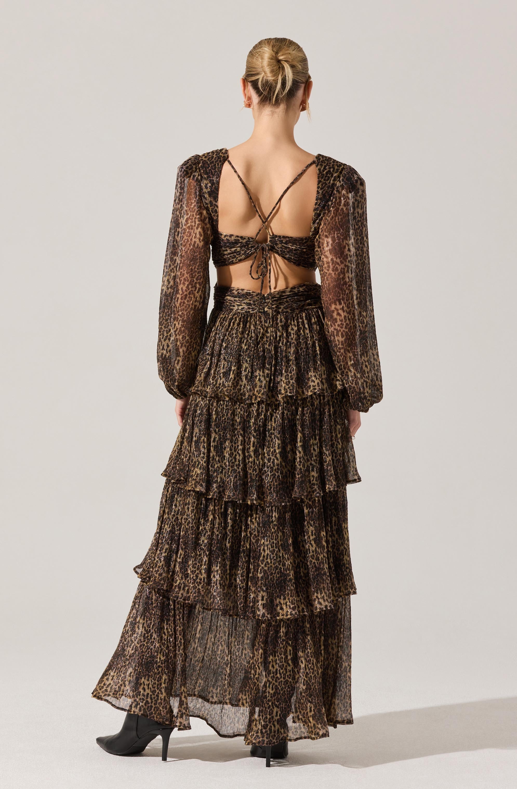 A woman is wearing a matching leopard print outfit with a long, tiered skirt and a crop top with long, semi-sheer puff sleeves. The crop top has thin spaghetti straps that crisscross at the back and ties at the front. The skirt has multiple ruffled layers and a high waist, creating a flowing silhouette. She is also wearing high-heeled boots.