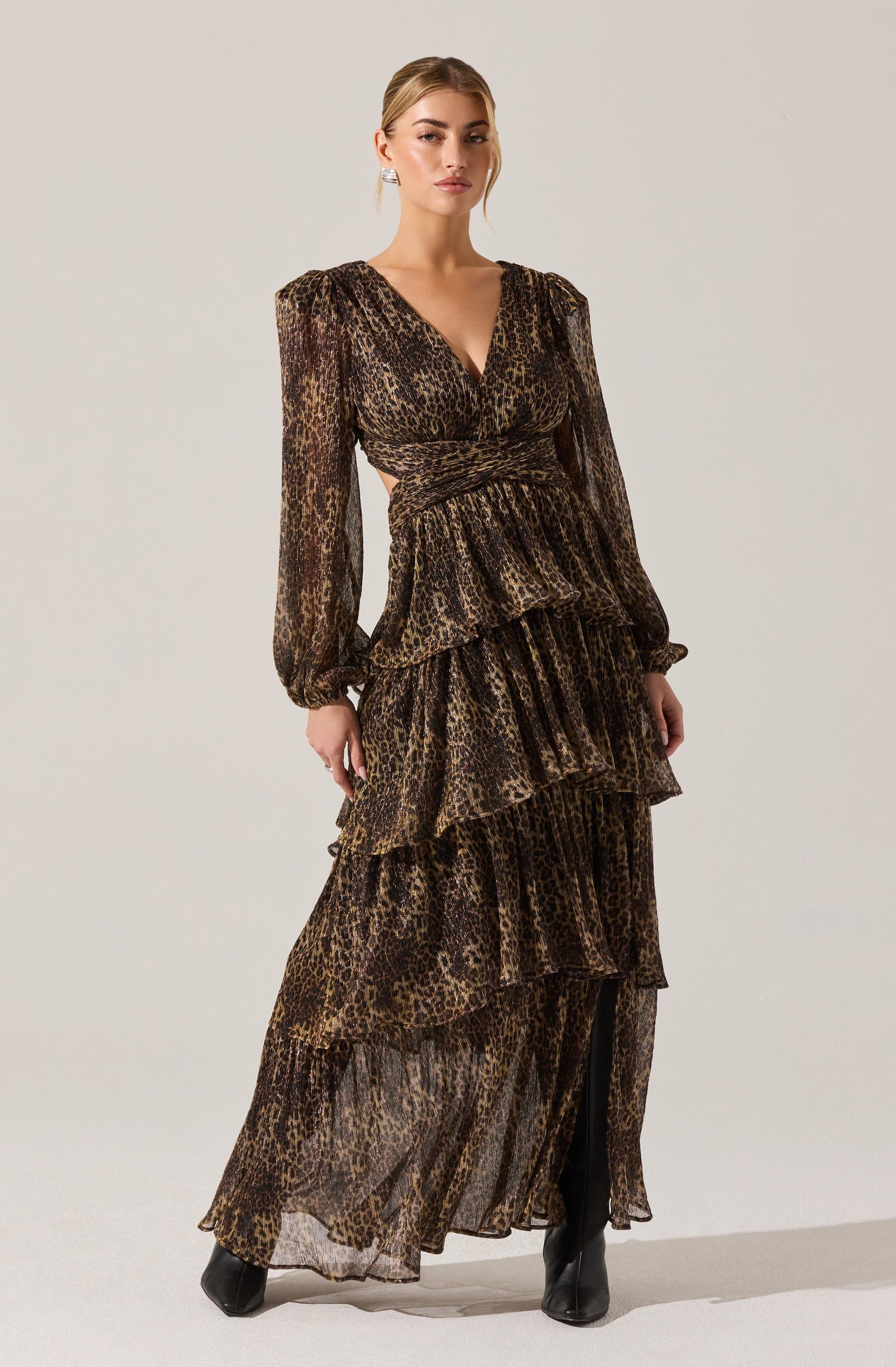 A woman is wearing a long, flowing dress with a leopard print pattern. The dress features long, semi-sheer sleeves with elastic cuffs, a V-neckline, and a fitted waist. The skirt has multiple tiered, ruffled layers that extend to the floor, creating a voluminous silhouette. She pairs the dress with black high-heeled boots.