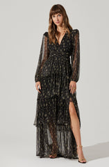 A woman is wearing a long, black, sheer dress with a floral pattern. The dress has puffy long sleeves, a V-neckline, and a fitted waist. It features multiple layered ruffles, with the skirt having a slit showing her leg. She is also wearing strappy gold high-heeled sandals.