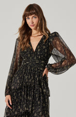 A woman wearing a black, semi-sheer dress with a sparkly, metallic floral pattern. The dress has a V-neckline, elastic waist, and long, billowy sleeves gathered at the wrists. Her hair is long and wavy with bangs, and she has a neutral expression.