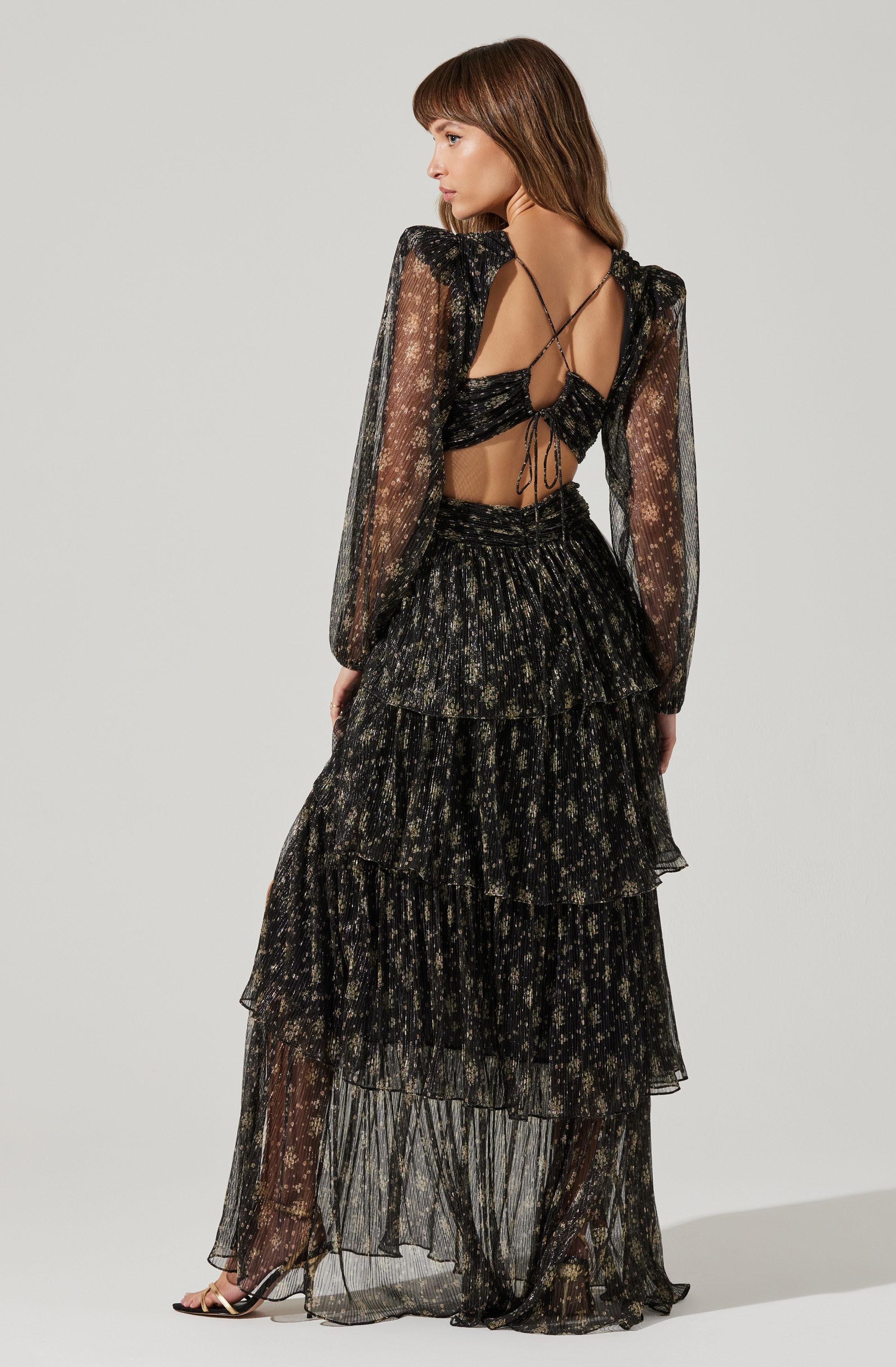 The outfit consists of a black, sheer, long-sleeved blouse with a delicate floral pattern, featuring a crisscross back design with ties. The matching high-waisted skirt is tiered with ruffled layers, also made of sheer fabric with a similar floral pattern, creating a coordinated, elegant look. The person is wearing high-heeled sandals with thin straps.