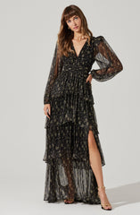A woman is wearing a long, black, sheer dress with a floral pattern. The dress features long, puffed sleeves, a deep V neckline, and a fitted waist. The skirt has multiple layered ruffles and a thigh-high slit. She is also wearing open-toed, high-heeled sandals.