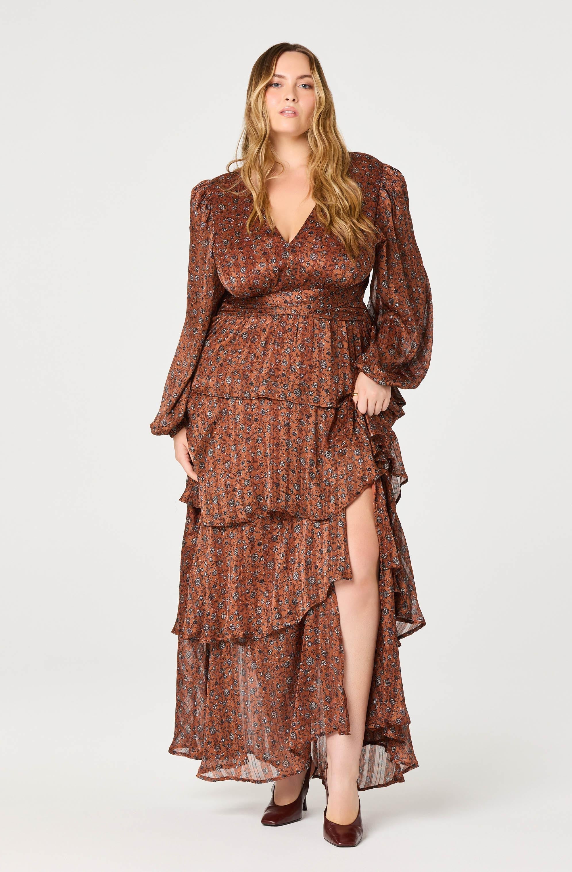 A woman is wearing a long, flowing dress with a rust-colored base and a small floral print pattern. The dress features a deep V-neckline and long, puffed sleeves gathered at the wrists. The waist is cinched, and the skirt has multiple layered ruffles with a high slit on the right side, revealing her leg. She completes the look with pointed-toe heels.
