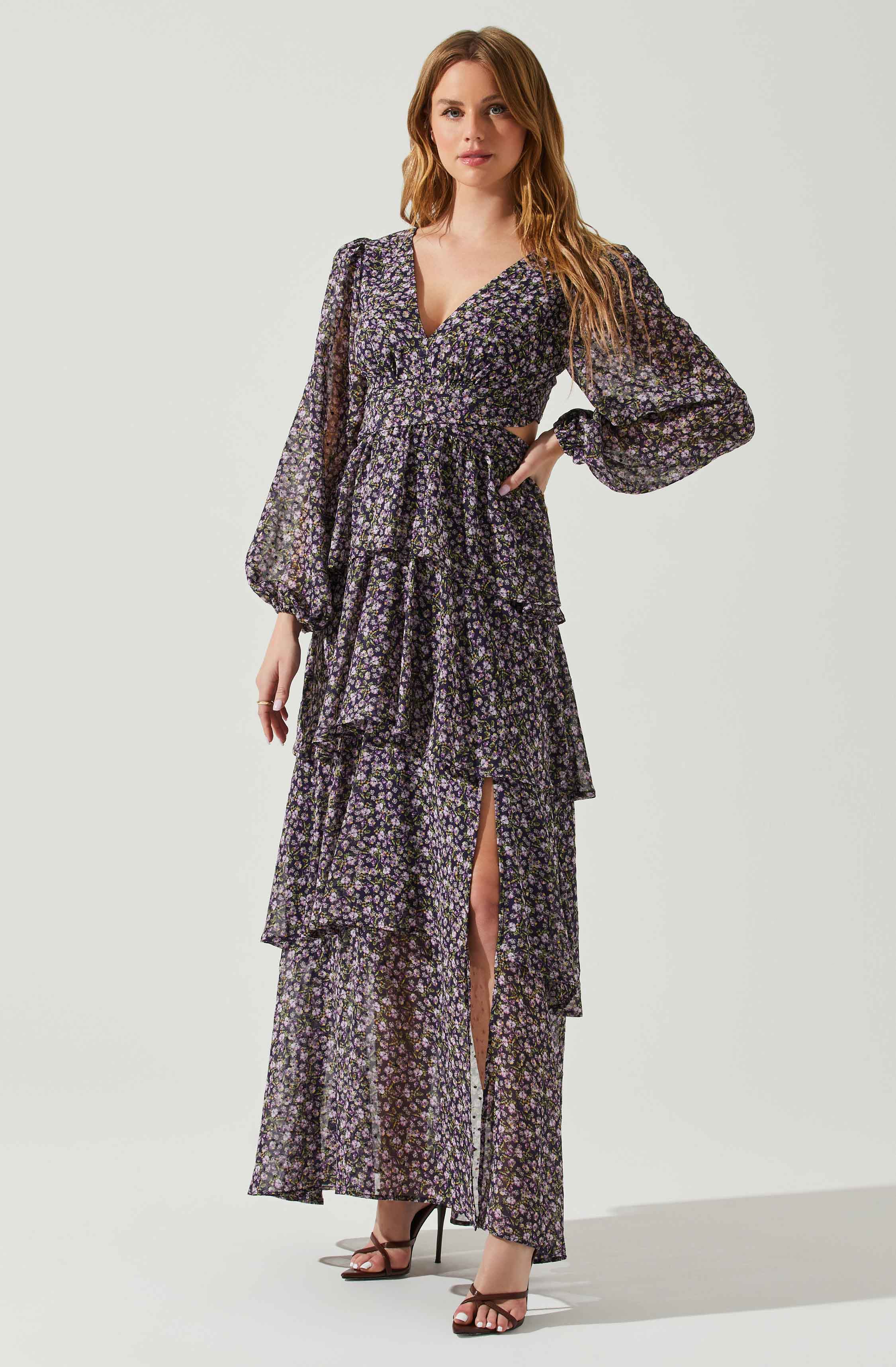 A woman is wearing a long, floral dress with small purple, pink, and white flowers on a black background. The dress features long, puffy sleeves with elastic at the cuffs, a deep v-neckline, and a fitted waist. The skirt has multiple layered tiers, with a thigh-high slit in the front revealing her legs. She is wearing high-heeled sandals with thin straps.