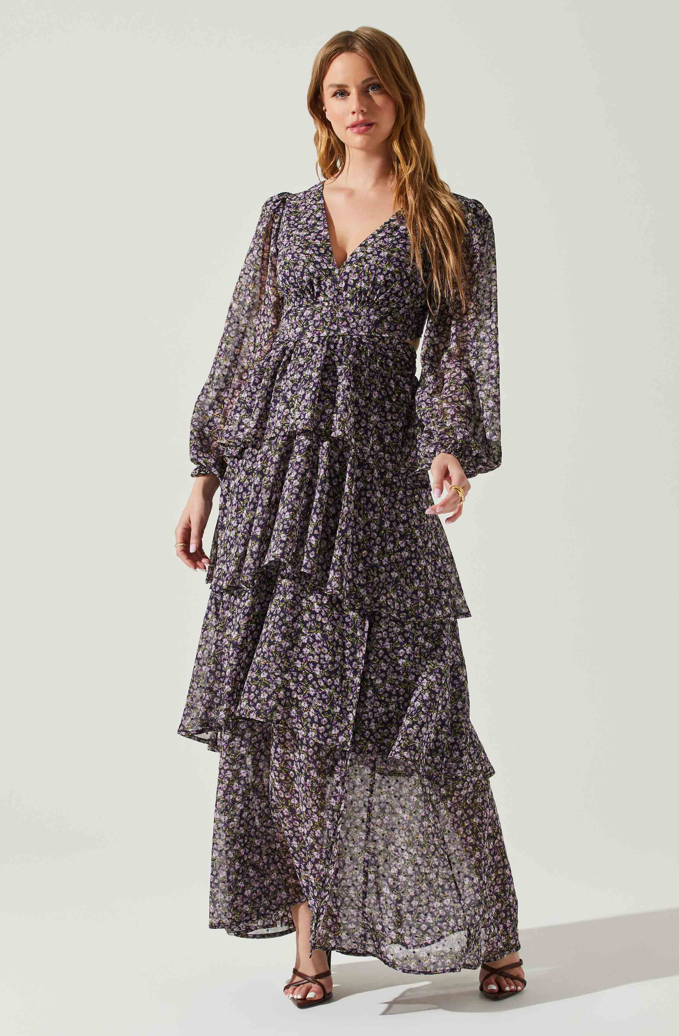 A woman is wearing a long, flowing dress made of sheer fabric with a small floral pattern in purple, white, and black. The dress features a V-neckline, long puffed sleeves with elastic cuffs, and a tiered, ruffled skirt that reaches the floor.