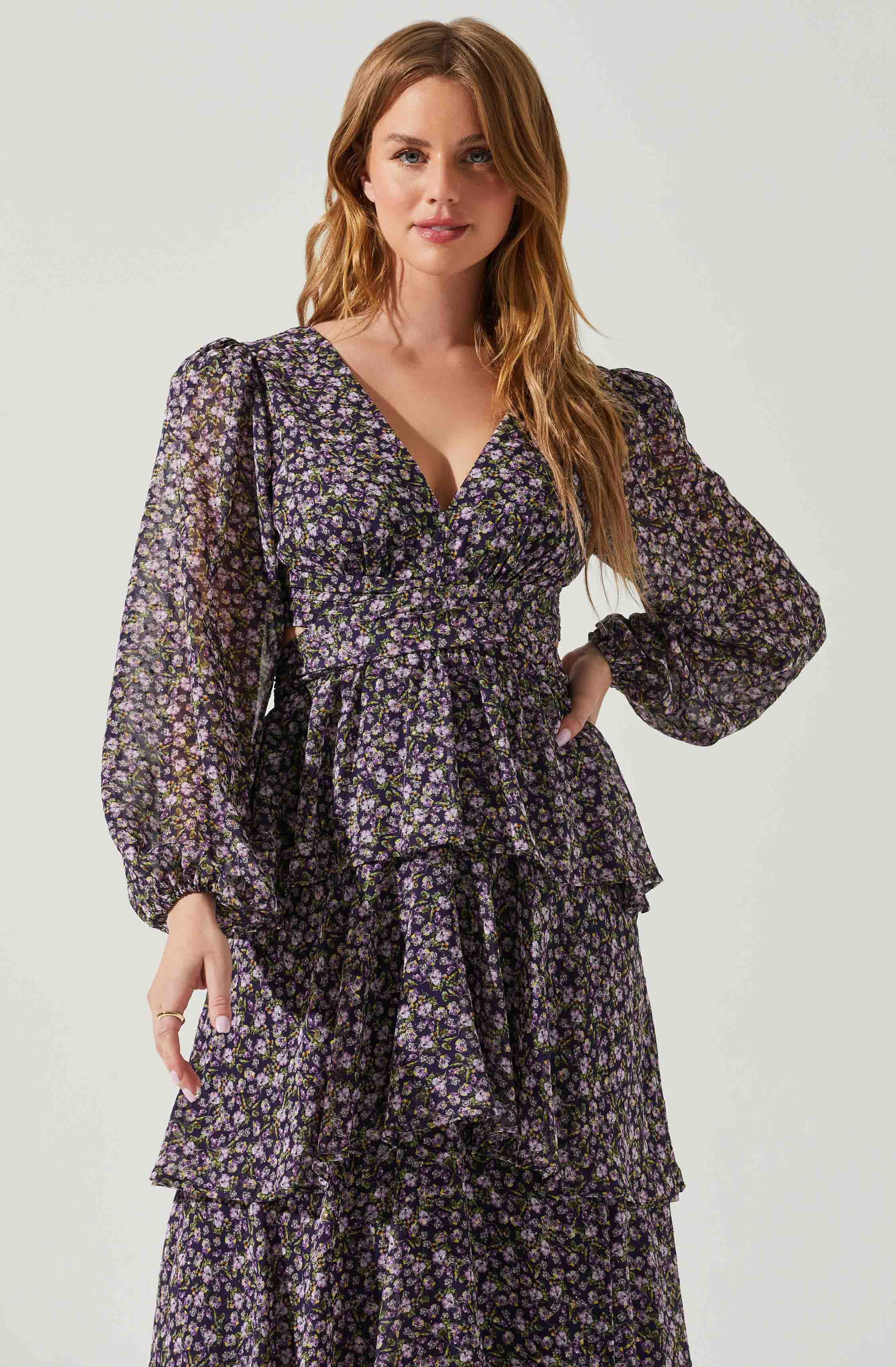 A woman is wearing a long, flowing dress with a dark background and a small floral pattern in shades of purple, pink, and white. The dress features long, semi-sheer puffed sleeves, a V-neckline, and a tiered, ruffled skirt.