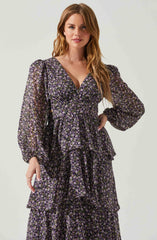 A woman is wearing a long, flowing dress with a dark background and a small floral pattern in shades of purple, pink, and white. The dress features long, semi-sheer puffed sleeves, a V-neckline, and a tiered, ruffled skirt.