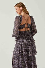 A woman is wearing a matching two-piece outfit made of sheer, black fabric with a small, purple and white floral print. The top has long, puffed sleeves and an open back with thin, crisscross straps and a tie detail at the lower back. The skirt is tiered with ruffle accents and a slit on the side.
