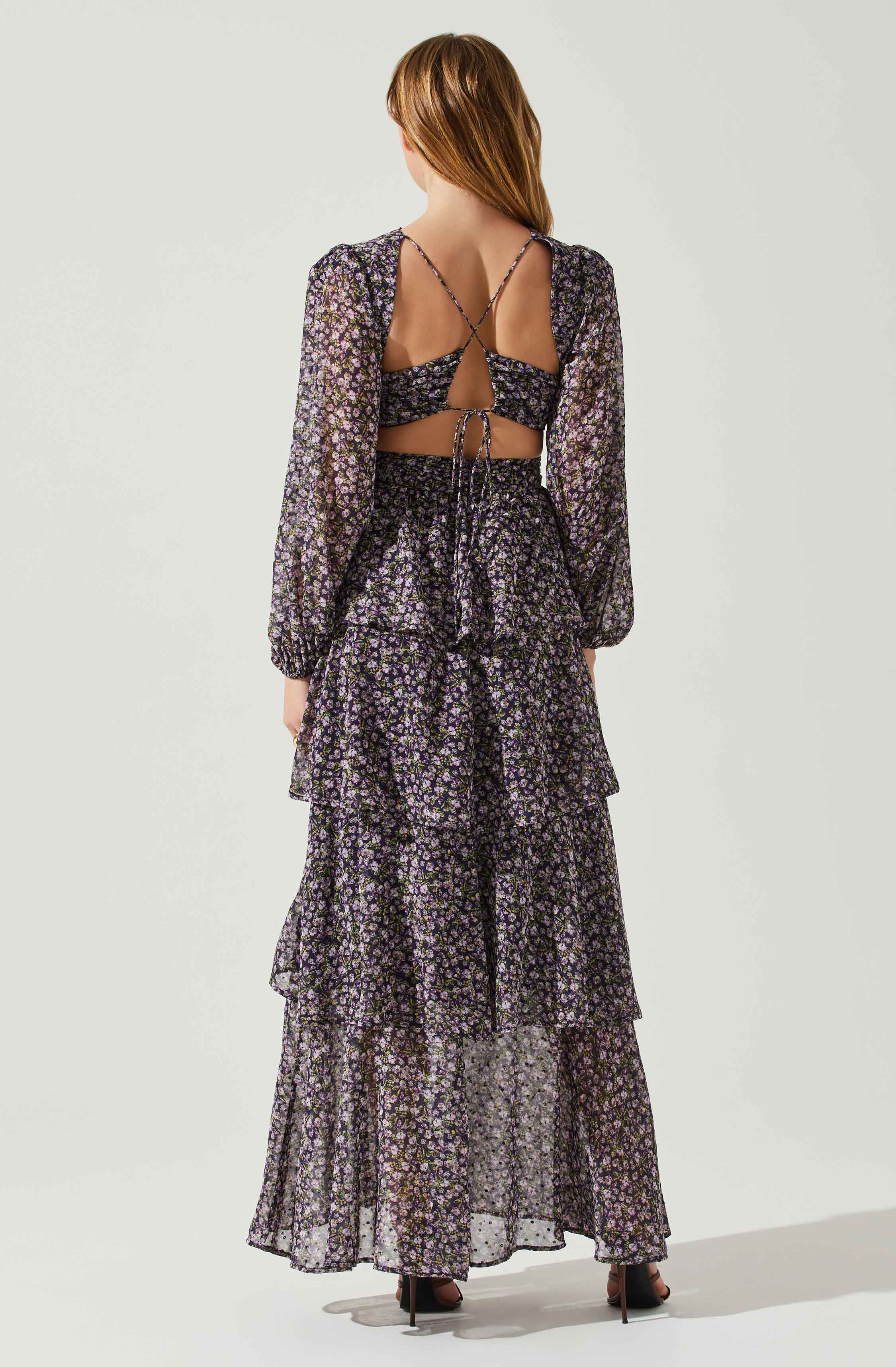 A woman is wearing a long, dark-colored dress with a floral pattern. The dress features multiple tiered, ruffled layers and has long, sheer sleeves. The back of the dress has a cut-out design with thin straps crisscrossing and tying at the waist.