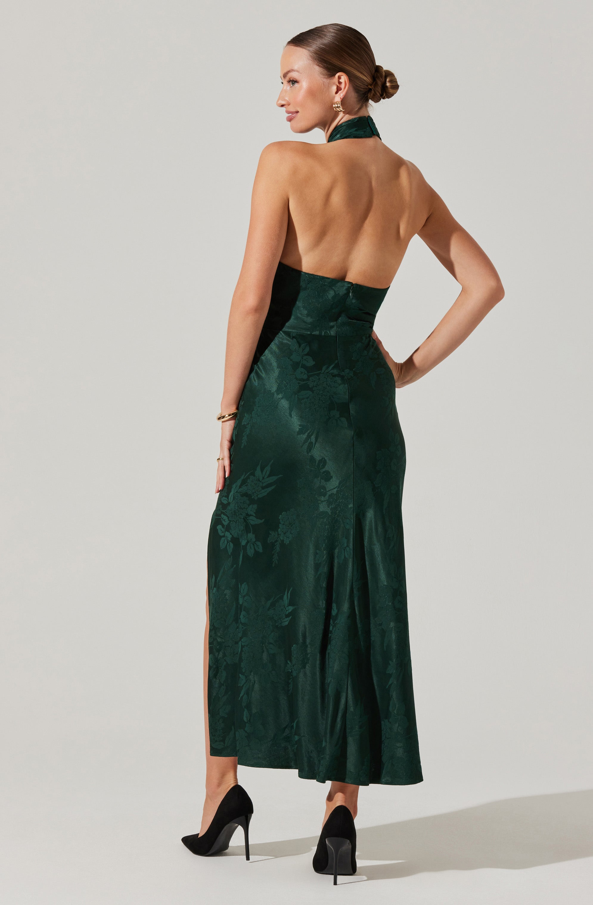 A woman is wearing a dark green, sleeveless evening gown with a halter neckline. The dress is fitted at the waist and features a subtle floral pattern with a slit up one side. She is also wearing black high-heeled shoes and gold jewelry, including earrings and a bracelet.