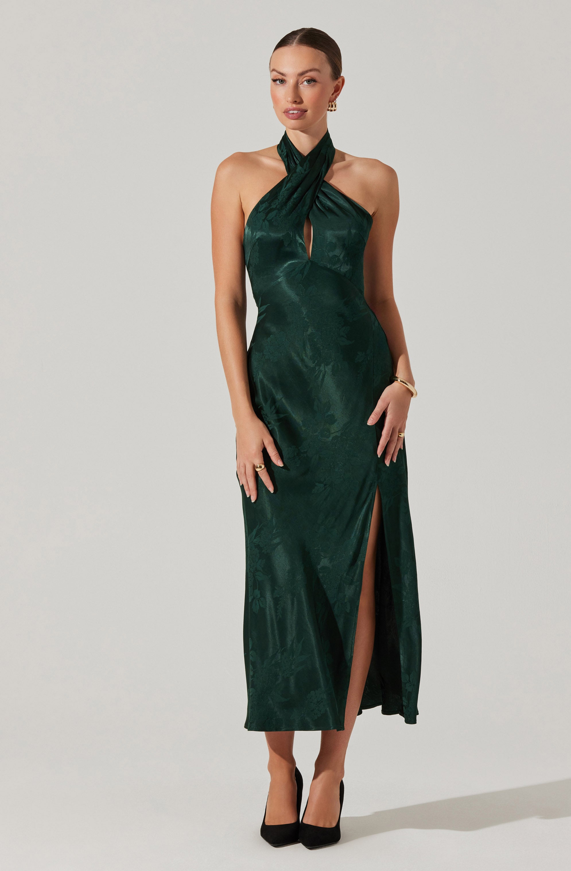 A woman is wearing a dark green satin halter neck evening gown with a textured pattern. The dress features a keyhole cutout at the chest and a high slit up one side, revealing her legs. She is accessorized with gold earrings, a bracelet, and rings, and is wearing black high-heeled shoes.